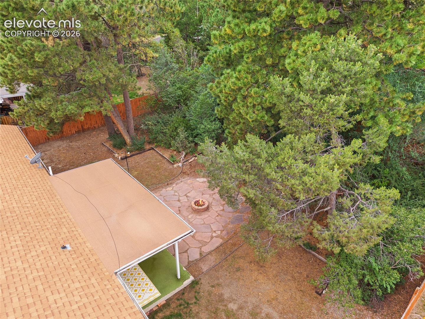 Backyard featuring a stone-paver patio, circular built-in fire pit, covered patio space, and mature trees enclosed by a wooden fence
