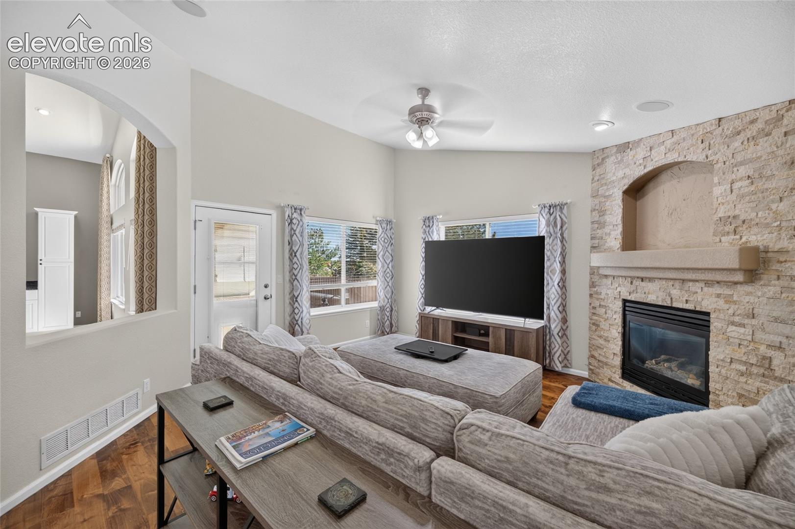 Living area featuring vaulted ceilings, stone-finish fireplace with built-in recessed niche, wood-finish flooring, and ceiling fan