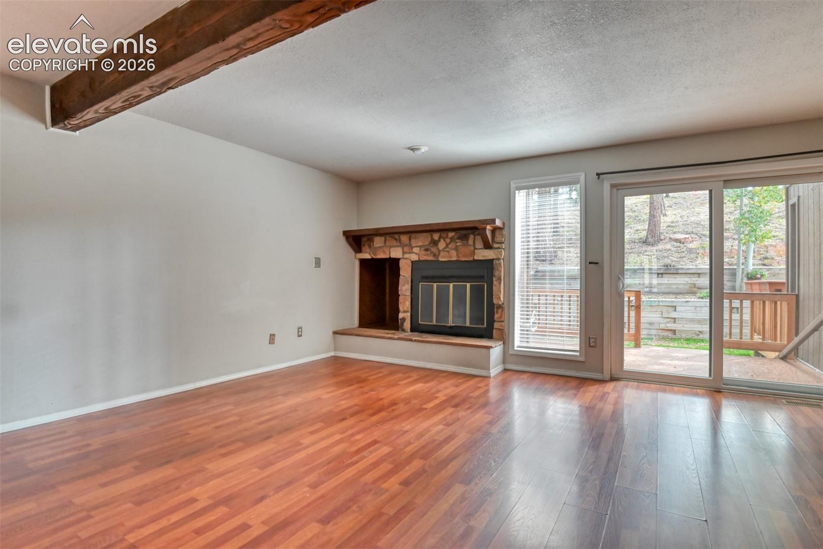 Wood-finish flooring throughout, stone-clad fireplace with wood mantel, exposed ceiling beam, sliding glass patio door, and an additional vertical window
