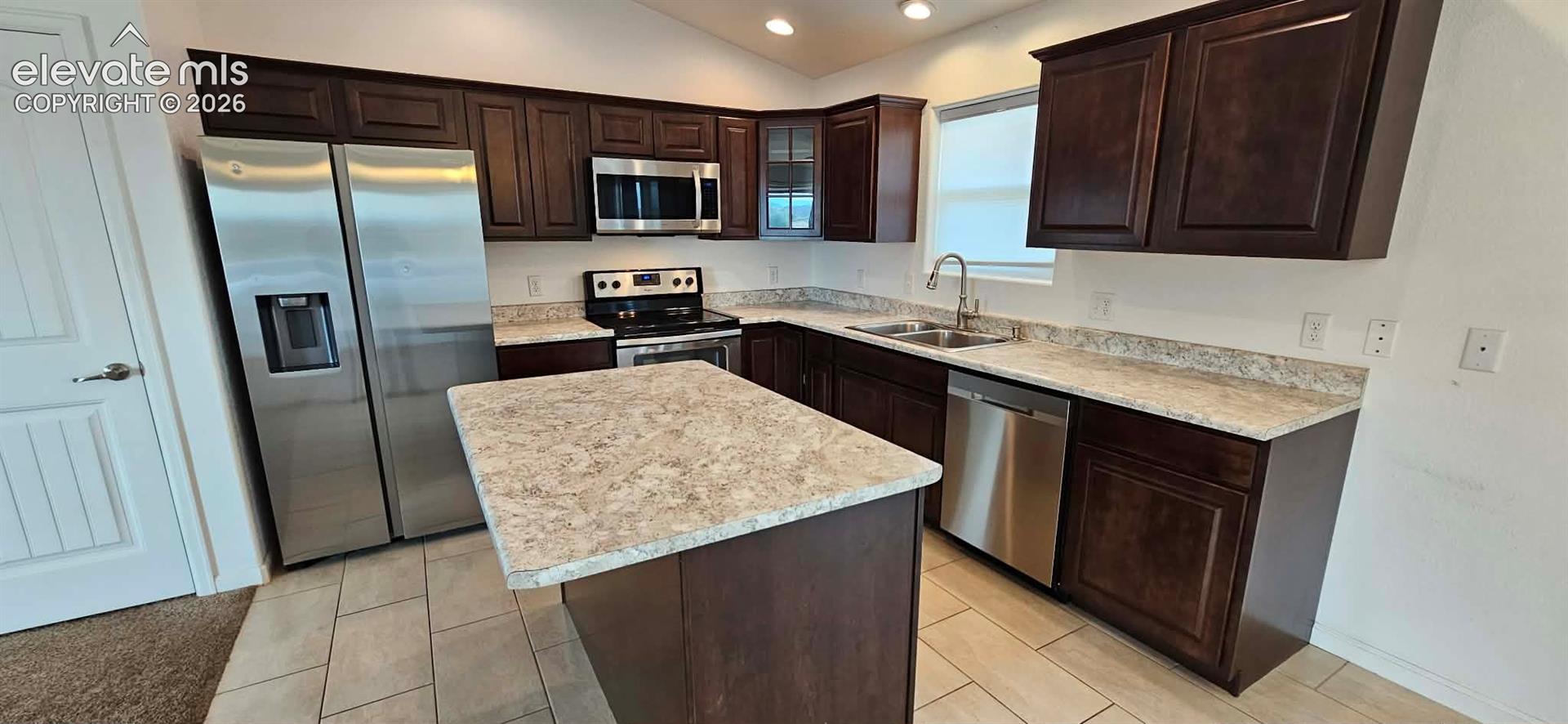 Kitchen featuring dark wood cabinetry, a central island, patterned stone-finish countertops, stainless steel appliances, and tiled flooring
