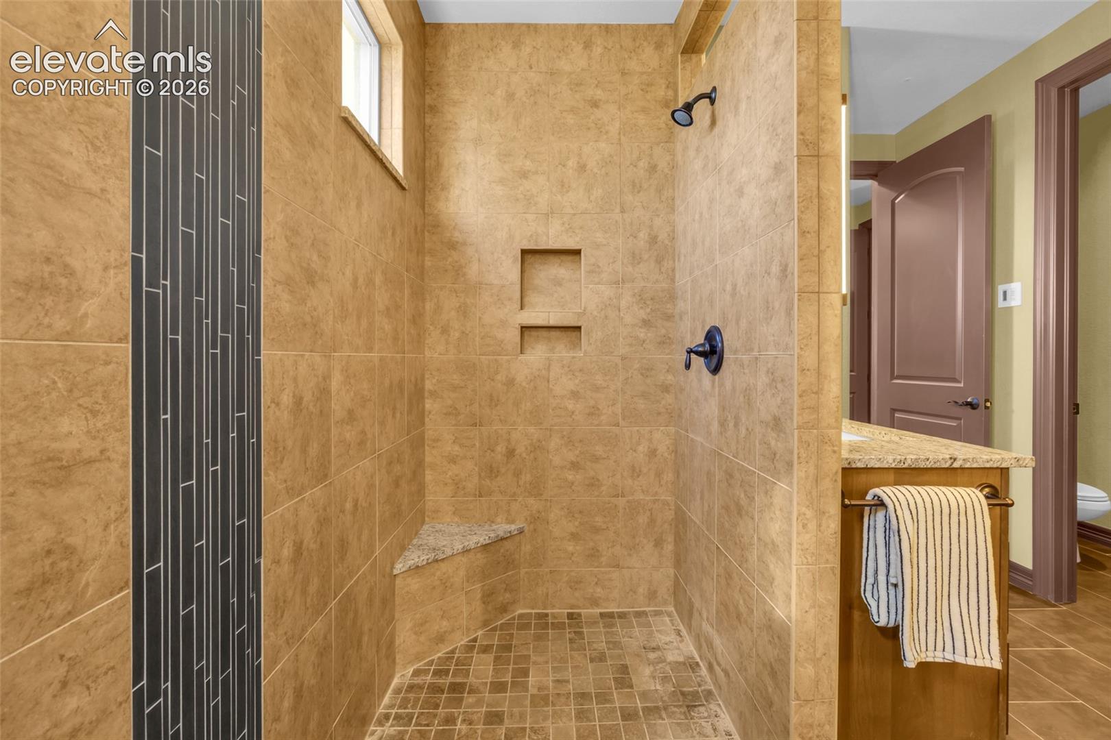 Walk-in shower featuring oversized earth-tone wall tile, vertical dark mosaic accent tiling, a stone-topped corner bench, dual recessed wall niches, and a dark-finish rainfall shower head