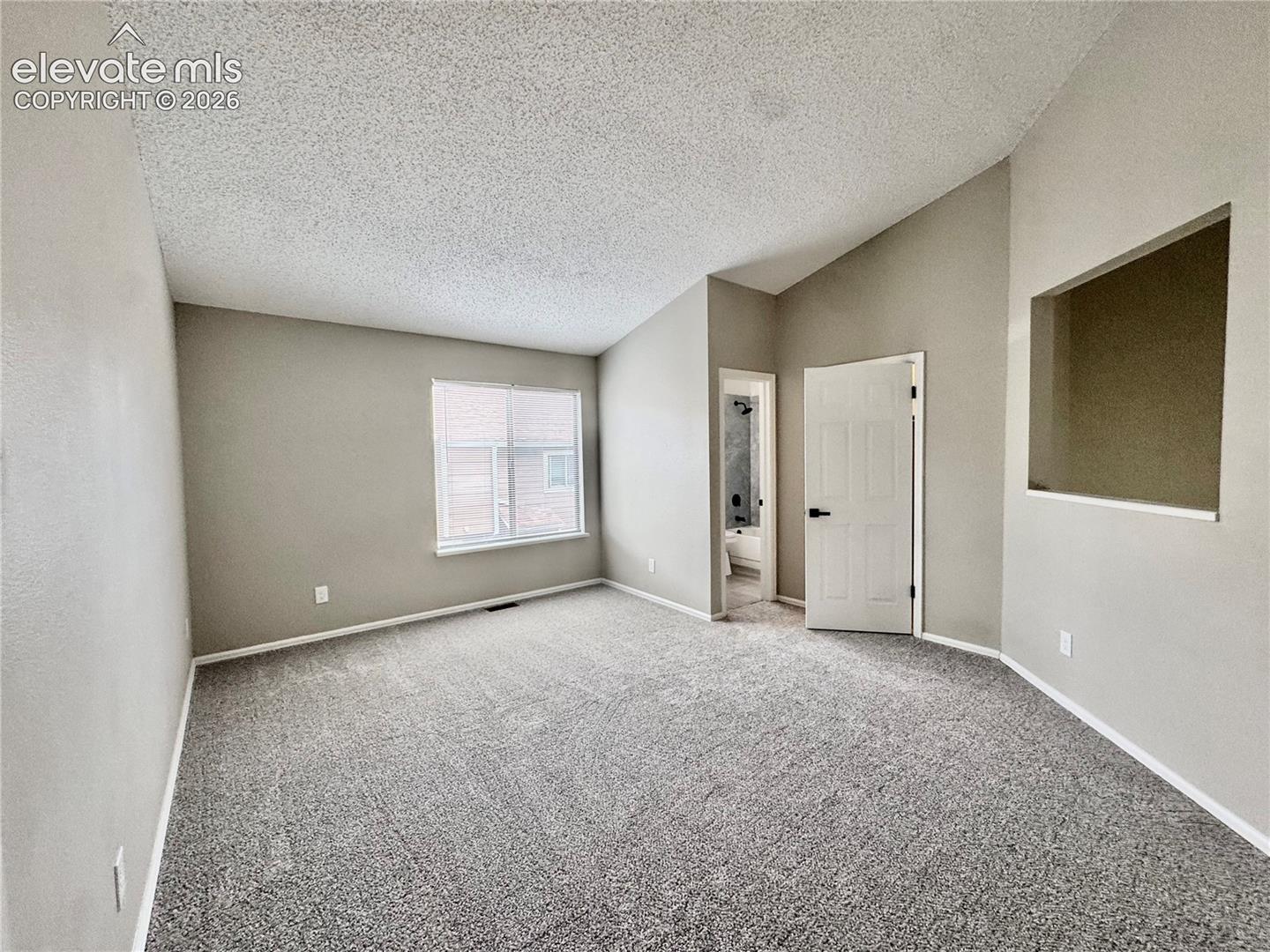 Vaulted ceiling with textured finish, neutral wall paint, gray carpeting, and a window with white trim