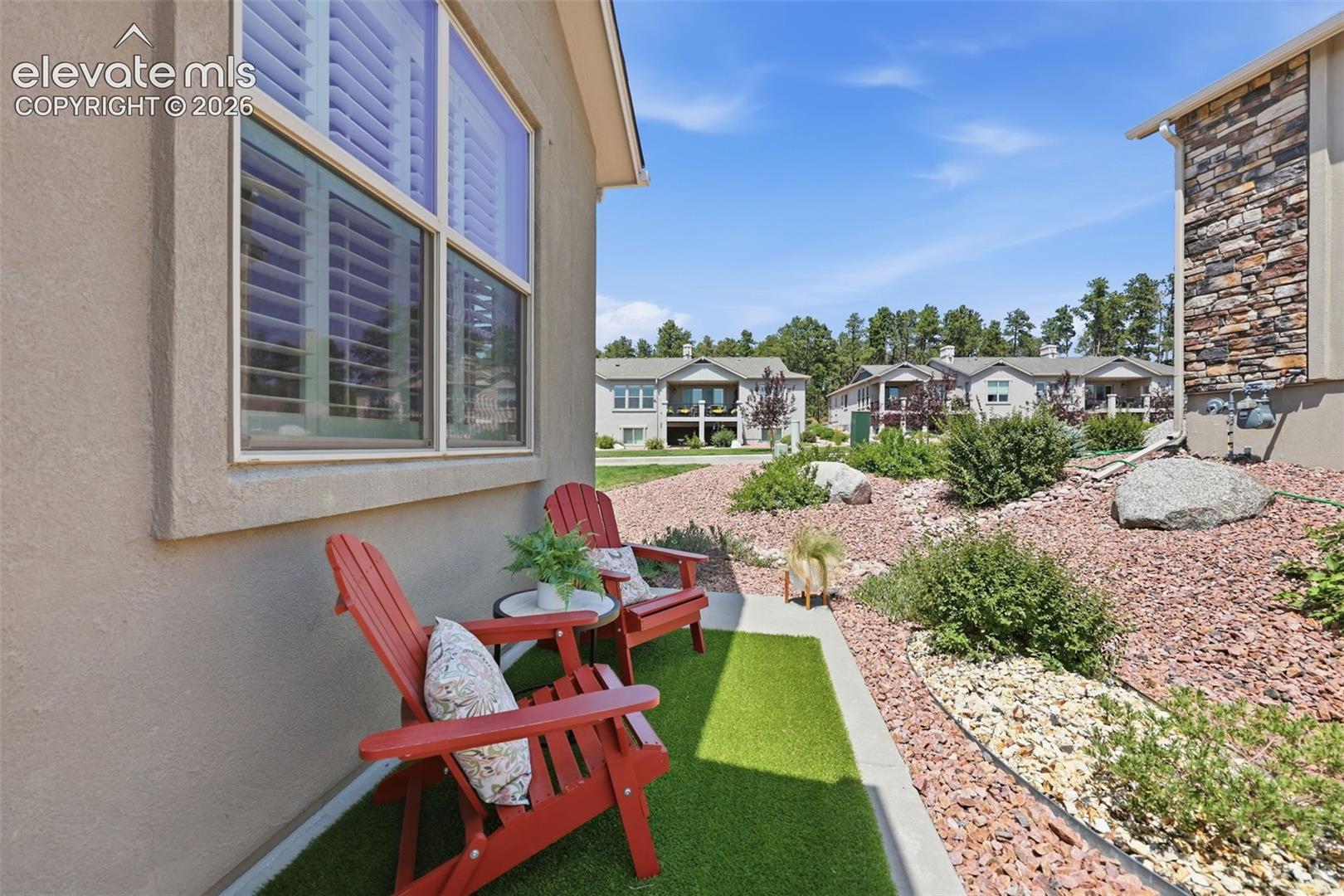 Exterior wall with stucco finish, large window with interior plantation shutters, and a compact outdoor patio featuring artificial turf.