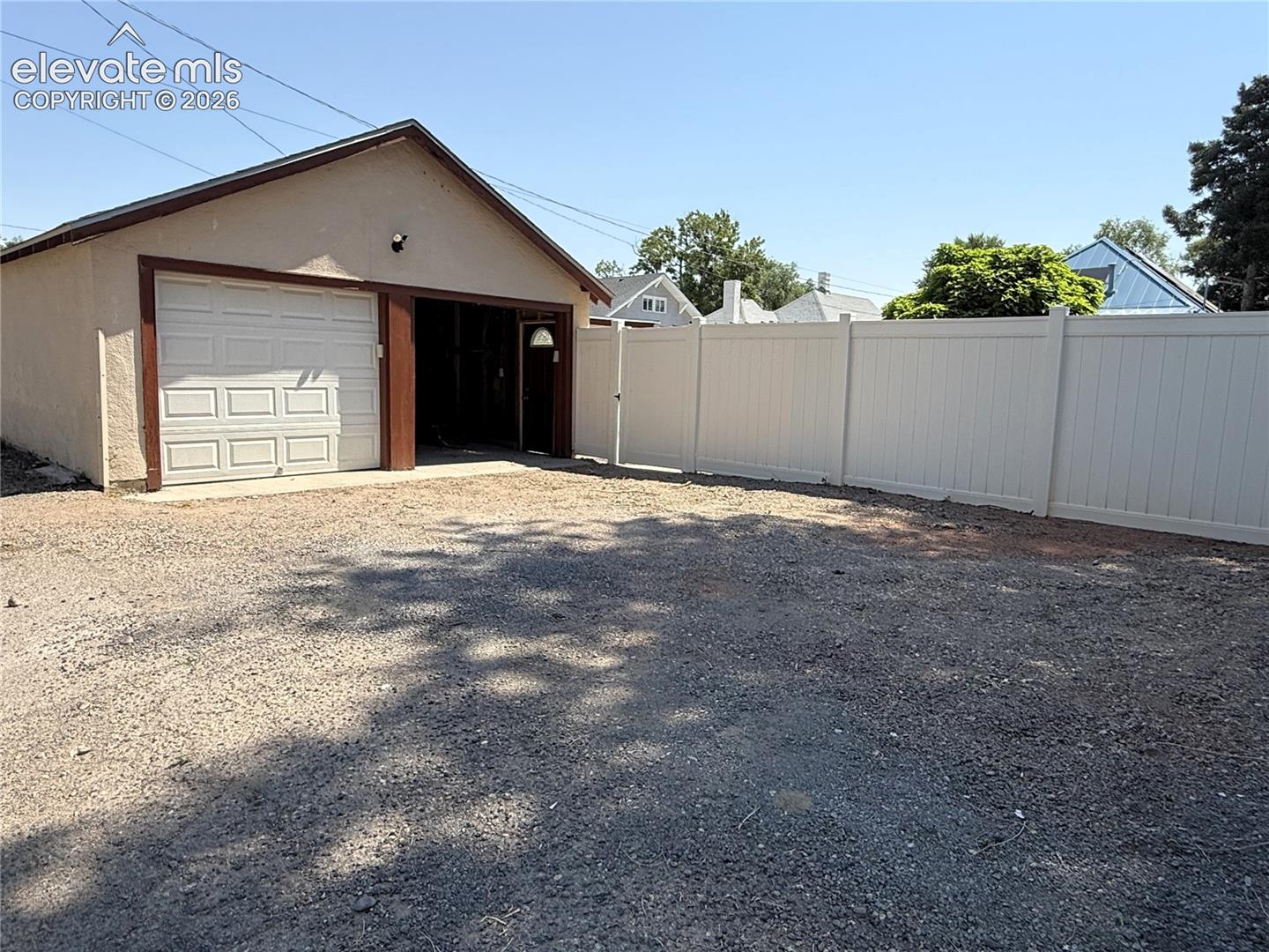 Detached garage featuring a single-car overhead door, service entry door, and gravel surfacing, enclosed by a white vinyl privacy fence