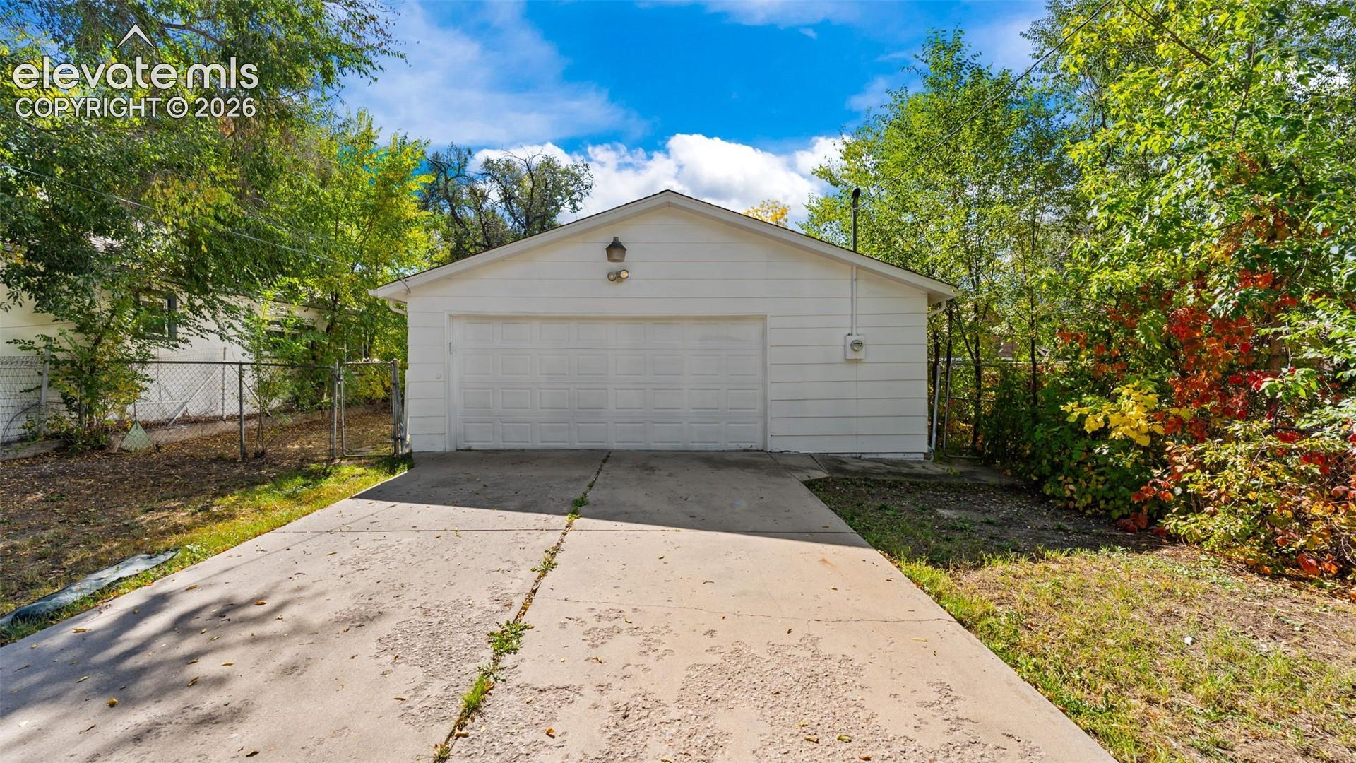 Spacious two-car garage with neutral siding exterior, concrete driveway, and mature landscaping with established trees and shrubs surrounding the property