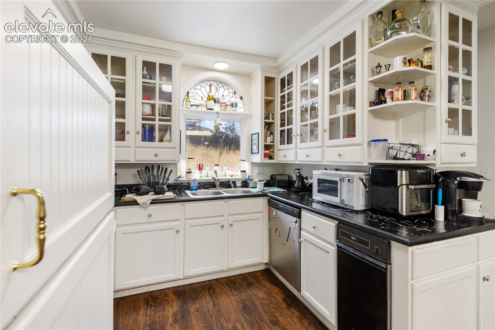 Kitchen featuring white shaker-style cabinetry, glass-paneled upper cabinets, dark stone-finish countertops, wood-finish flooring, and a decorative arched window above the dual-basin sink
