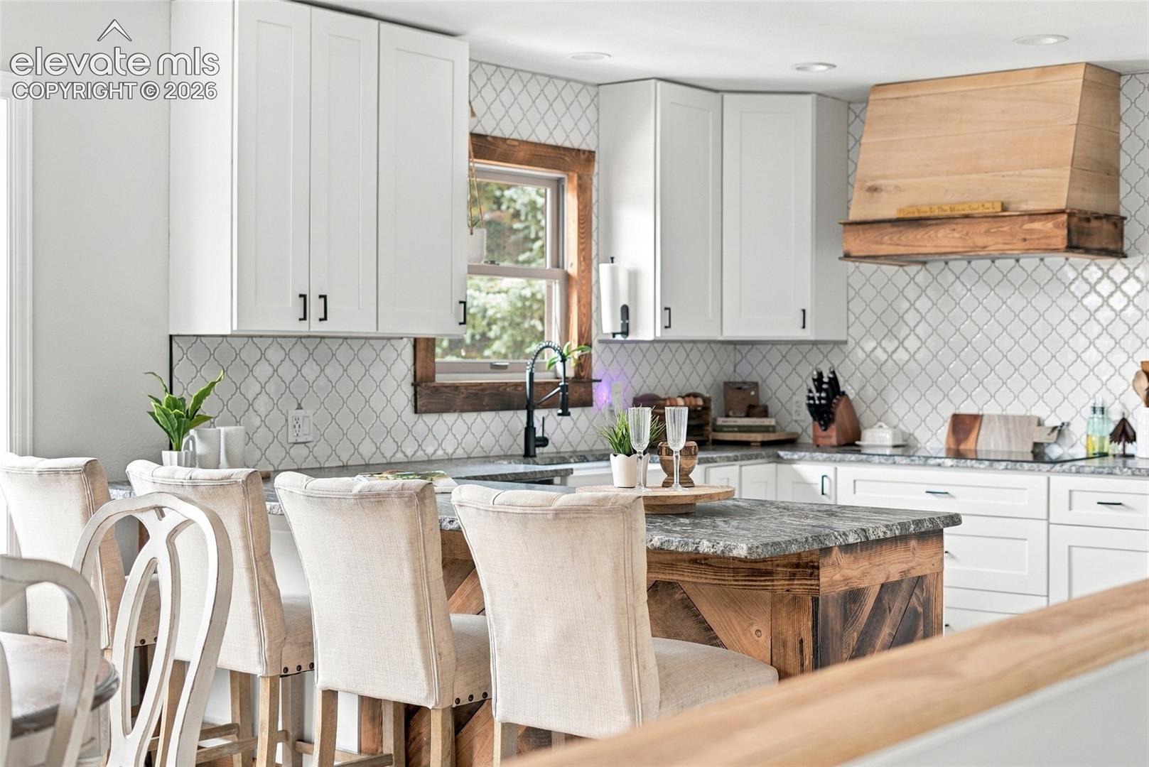 White shaker-style cabinetry with matte black hardware, textured tile backsplash, wood-finish range hood, and a kitchen island with a stone countertop