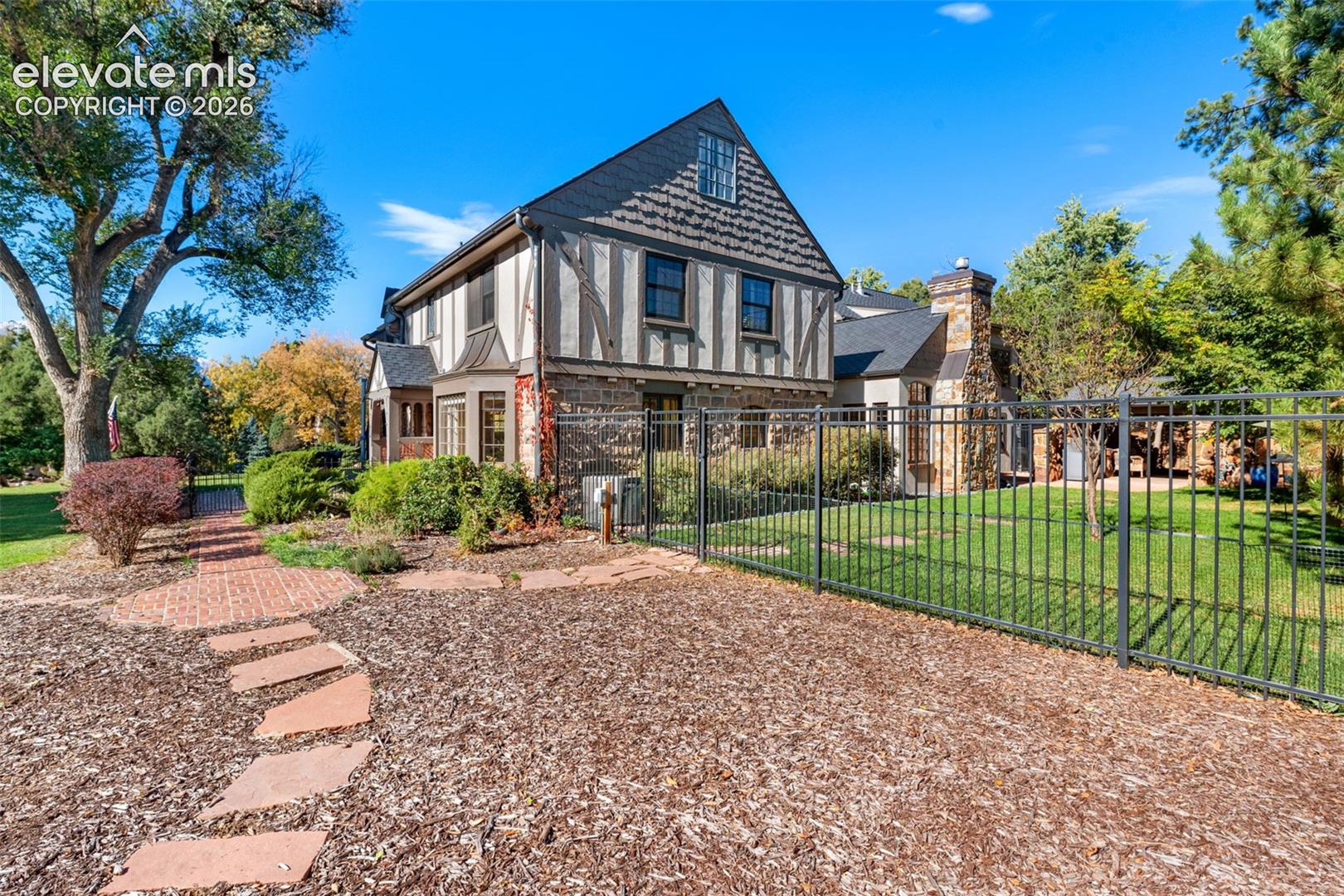 Tudor-style exterior featuring half-timbering, stone veneer, slate-style roofing, a prominent stone chimney, and a decorative wrought-iron perimeter fence