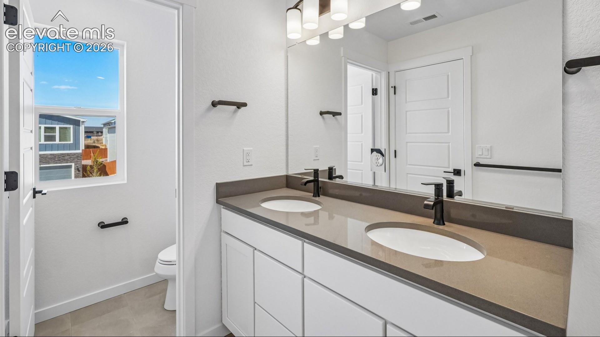 Dual-sink vanity with a solid-surface countertop, matte black fixtures, full-width mirror, and neutral tile flooring
