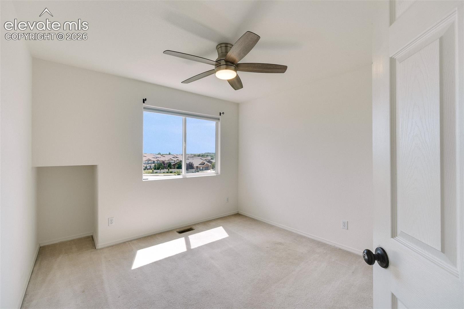 Bright interior with neutral carpet flooring, a ceiling fan, and a window offering a distant neighborhood scene