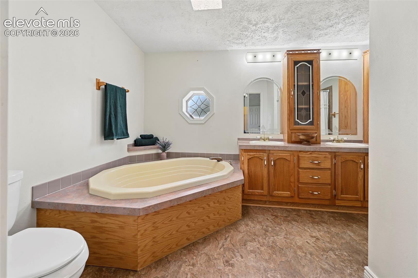 Built-in corner tub with surrounding countertop, dual vanity with wood-finish cabinetry, arched mirrors, and an octagonal decorative window