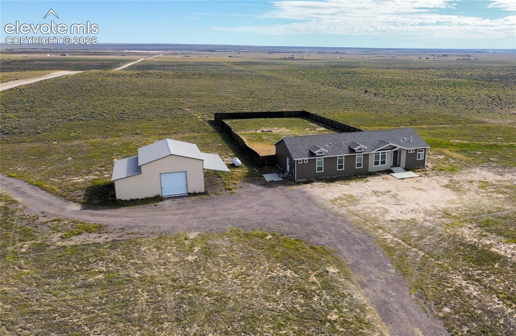 Single-story residence featuring a shingled roof and dark siding, accompanied by a detached metal-sided outbuilding, a fenced enclosure, and an expansive gravel driveway