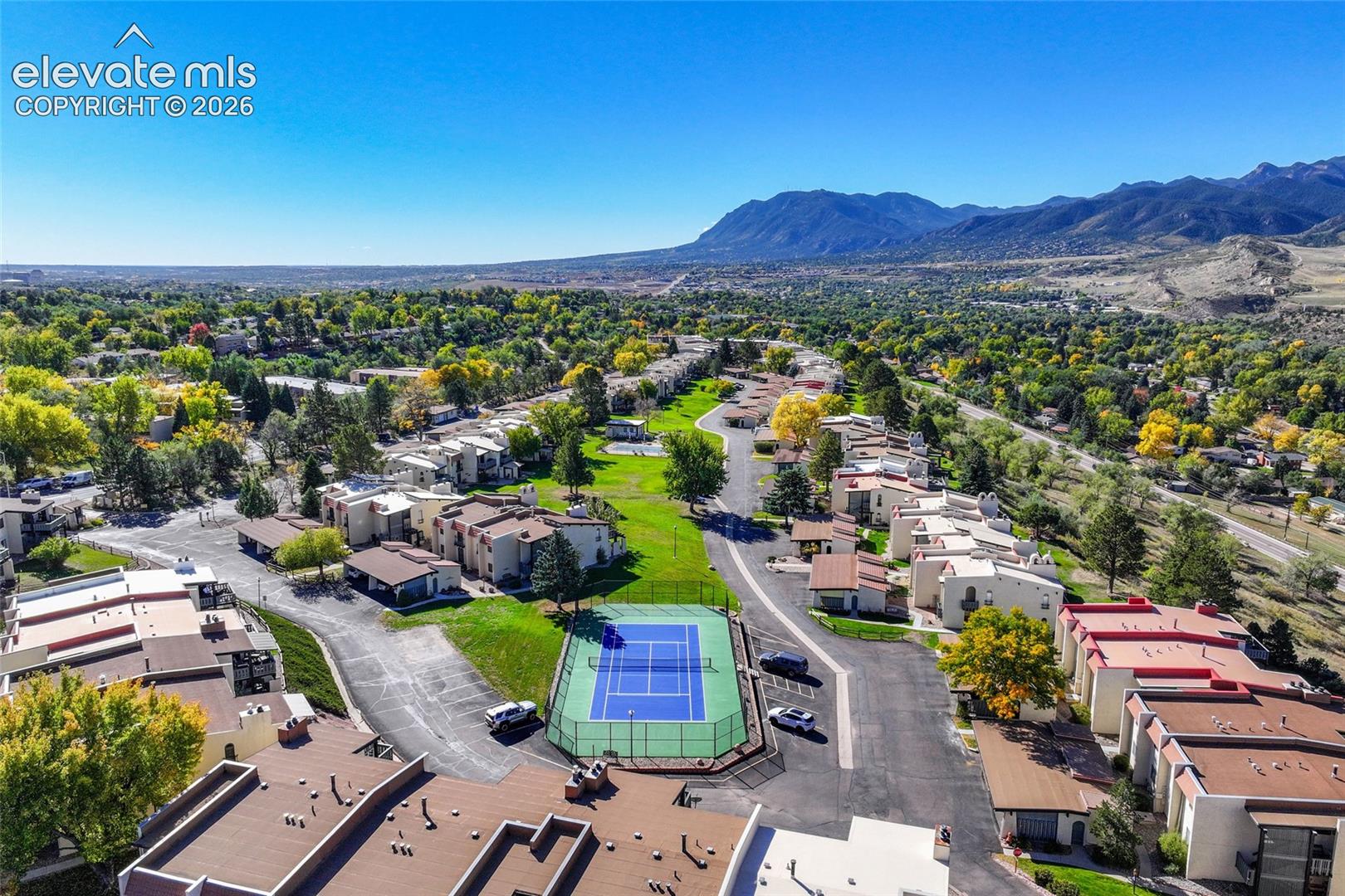 Aerial perspective of community grounds featuring a fenced tennis court, paved roadways, mature landscaping, and prominent mountain backdrop