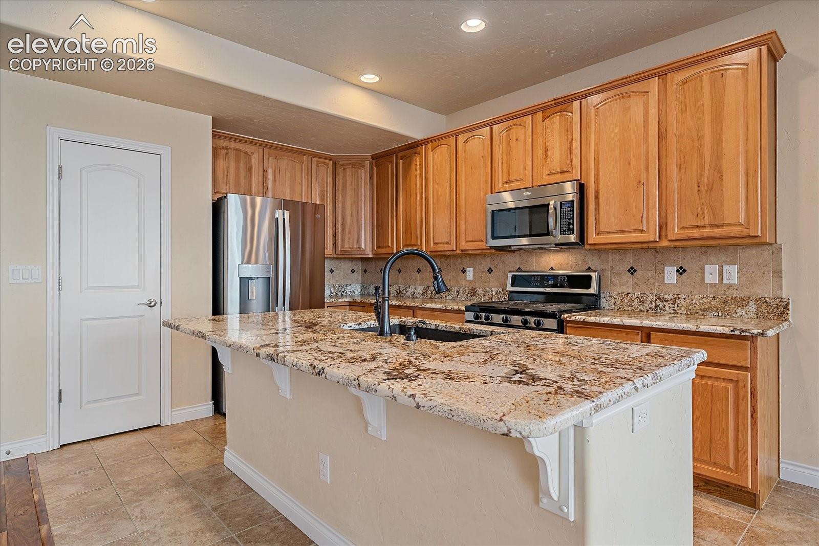 Kitchen featuring stained wood cabinetry, stone countertops, central island with a deep-basin sink, and stainless steel appliances