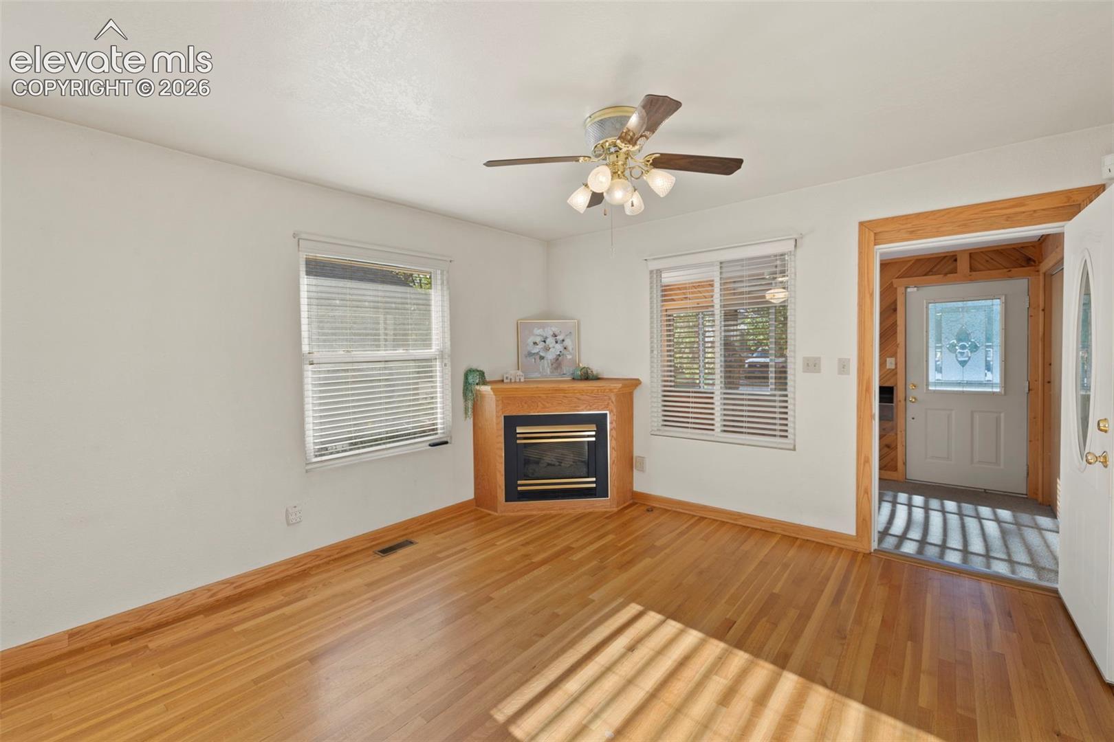 Wood-finish flooring throughout, corner fireplace, windows with blinds, ceiling fan with integrated lighting, and an entry door with decorative glass