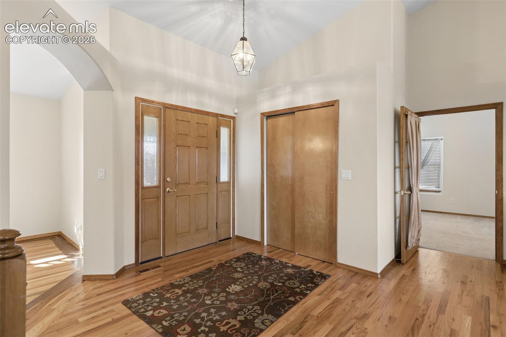 Foyer featuring wood-finish flooring, vaulted ceilings, a wood-paneled front door with sidelights, a hanging pendant light, and a built-in closet with sliding wood doors