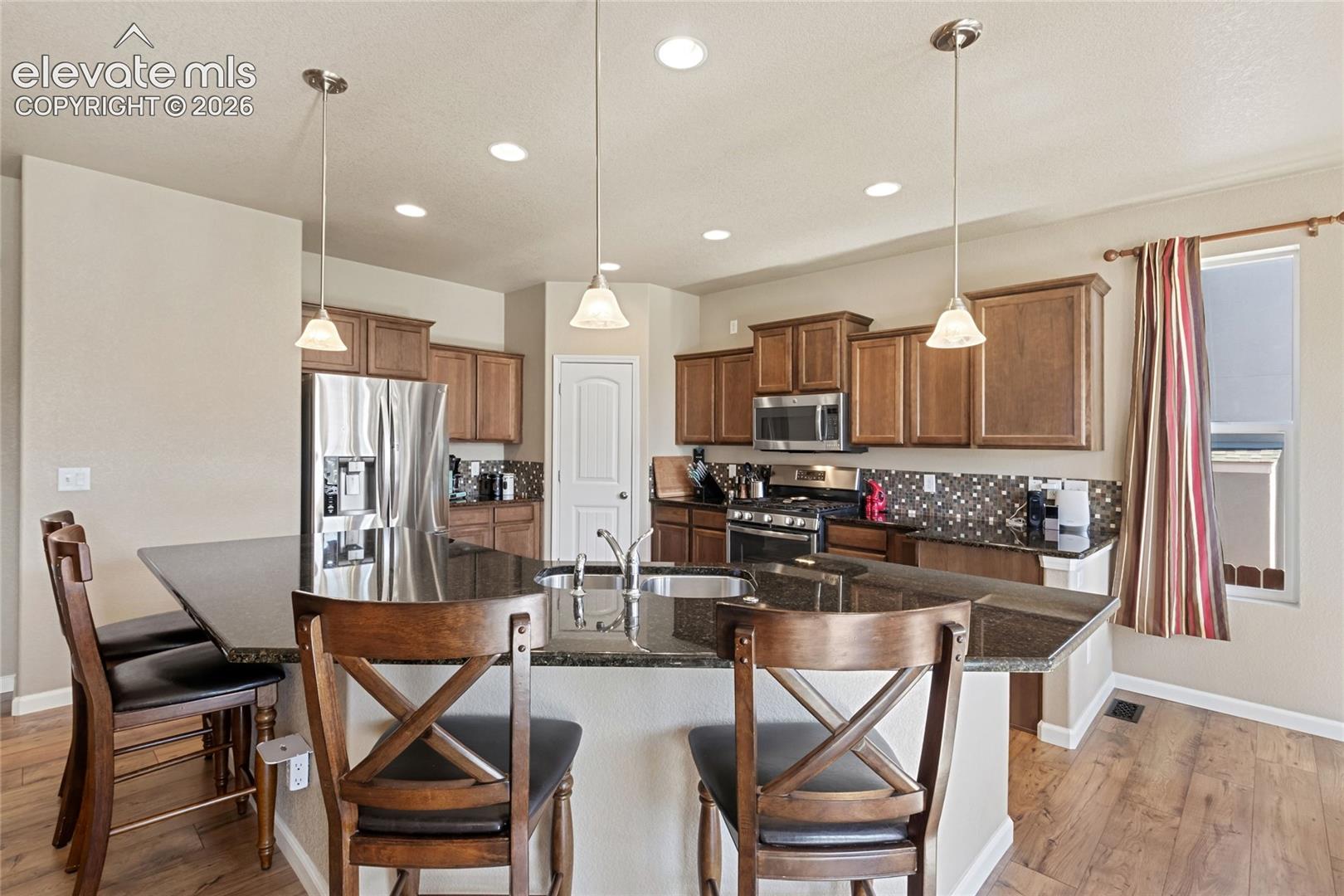 Kitchen featuring dark stone countertops, medium-tone wood cabinetry, stainless steel appliances, a center island with integrated sink, and recessed lighting