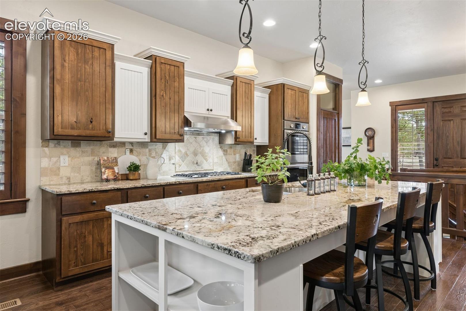 Kitchen featuring a large central island with stone-finish countertops, custom two-tone cabinetry, professional-grade stainless steel wall oven, decorative tile backsplash, and hanging pendant light fixtures