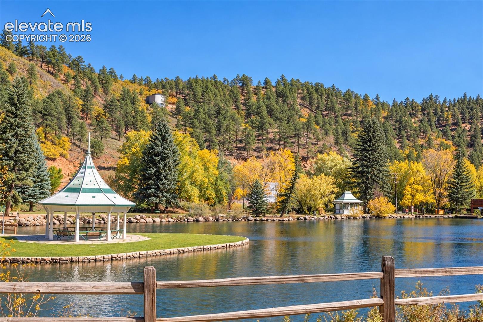 Waterfront parcel featuring a wooden split-rail fence, a manicured lawn, a white-framed gazebo with a conical roof, and a stone-lined shoreline