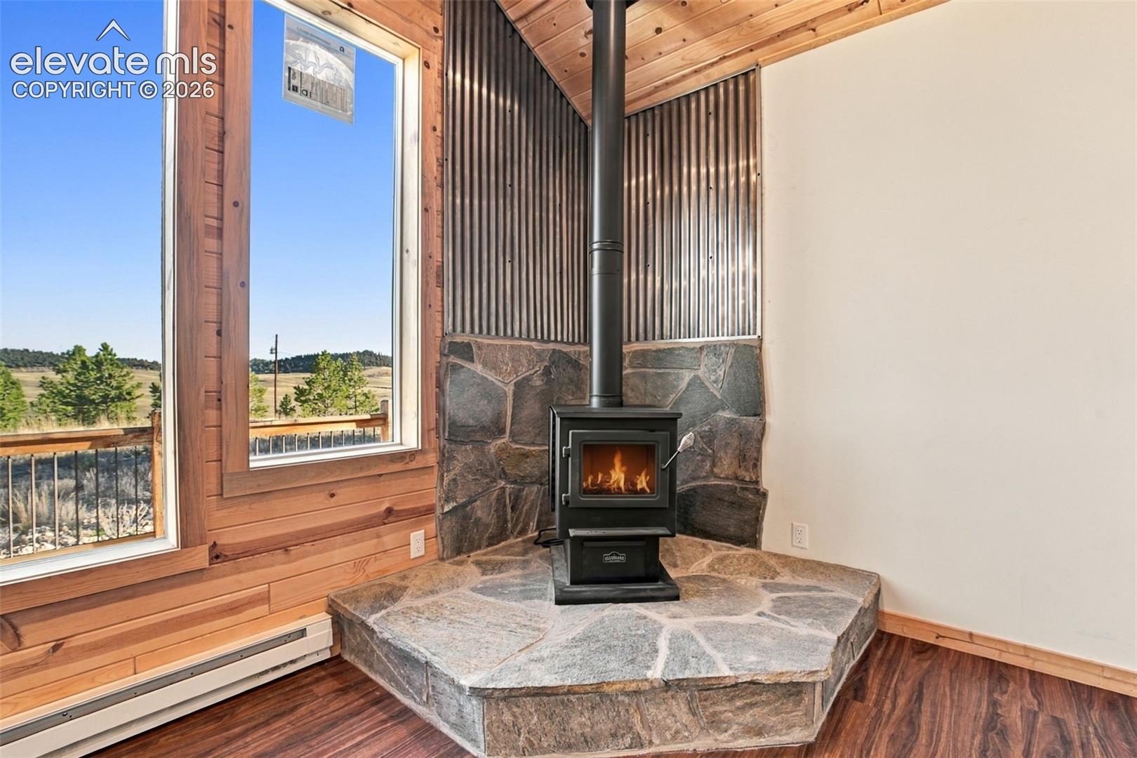 Corner wood-burning stove on a raised stone hearth, featuring a metal-paneled wall surround, wood-plank walls and ceiling, and large picture windows