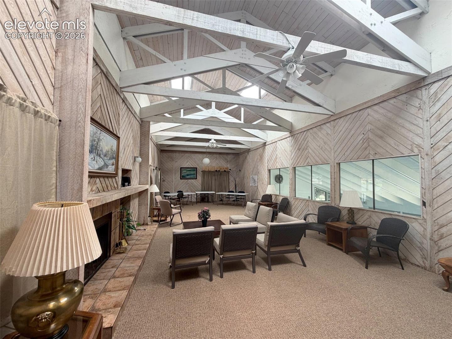 Vaulted ceiling with exposed white-washed wooden trusses, herringbone-patterned wood wall paneling, tile-surround fireplace, and large horizontal windows