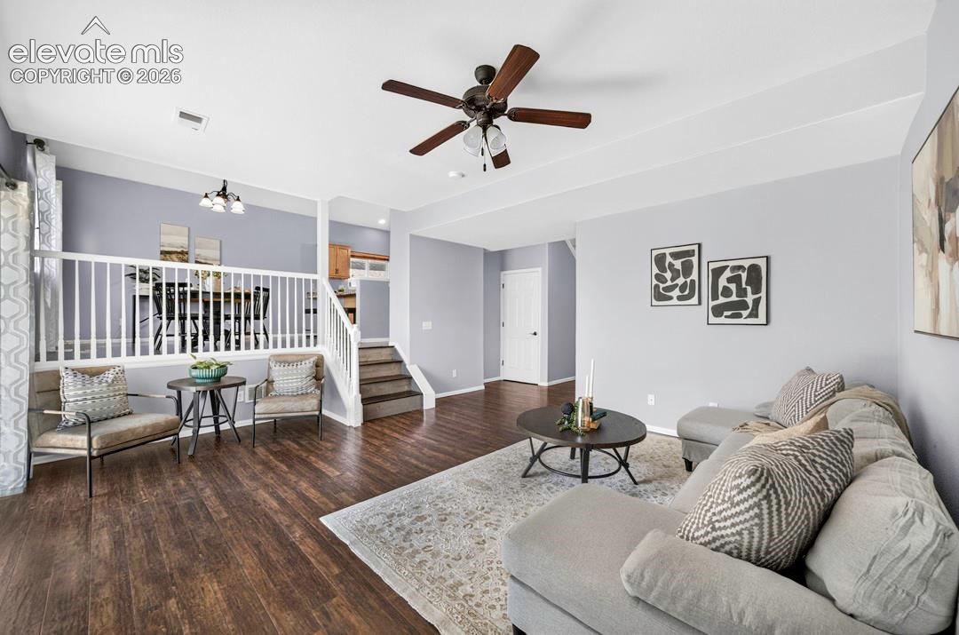 Open-concept interior featuring wood-finish flooring, tray ceiling, built-in wooden staircase railing, and a multi-blade ceiling fan with integrated light fixture