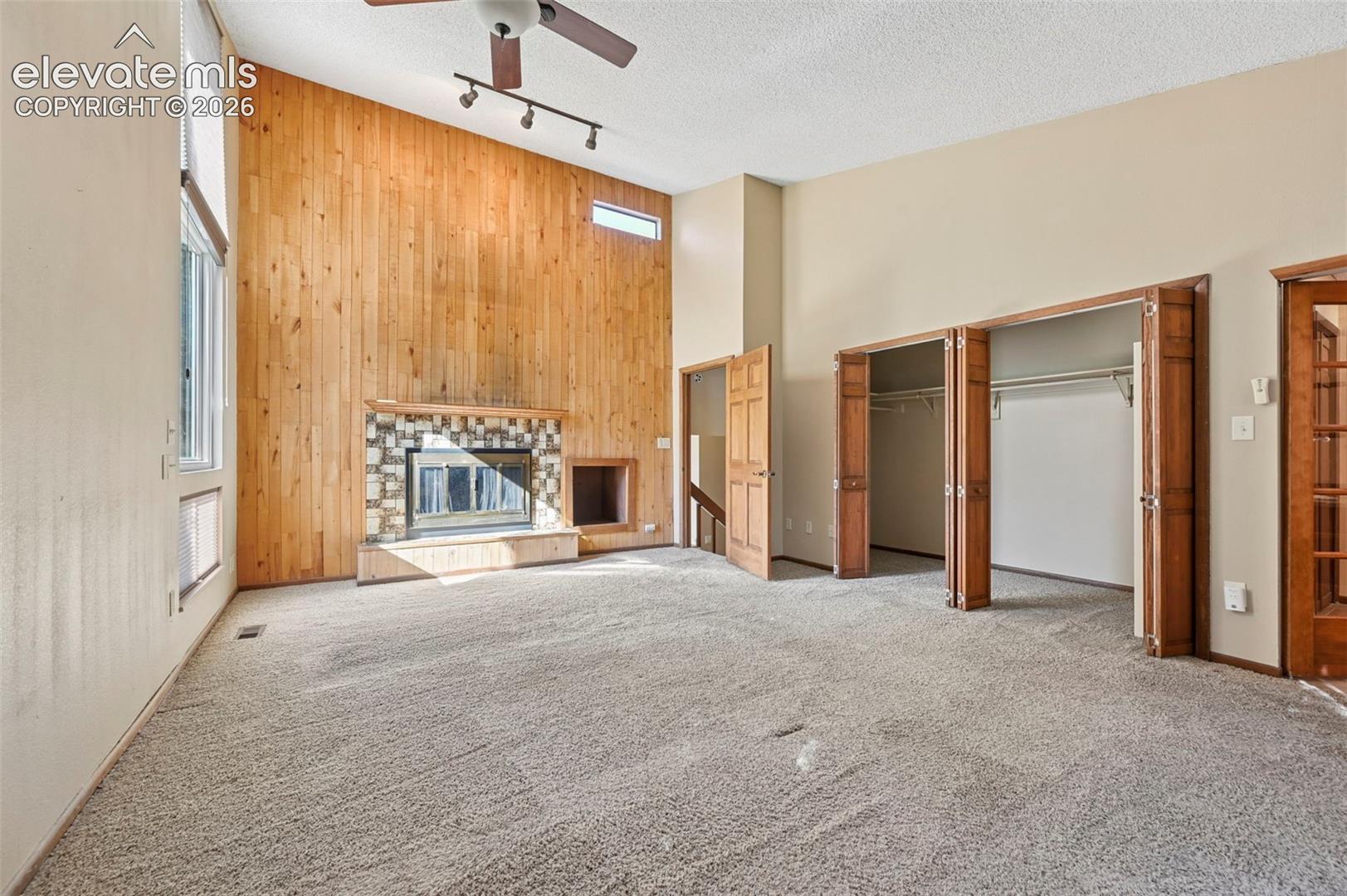 Vaulted ceiling featuring a wood-plank accent wall, a floor-to-ceiling stone-faced fireplace, integrated ceiling fan and track lighting, and a dual-door reach-in closet