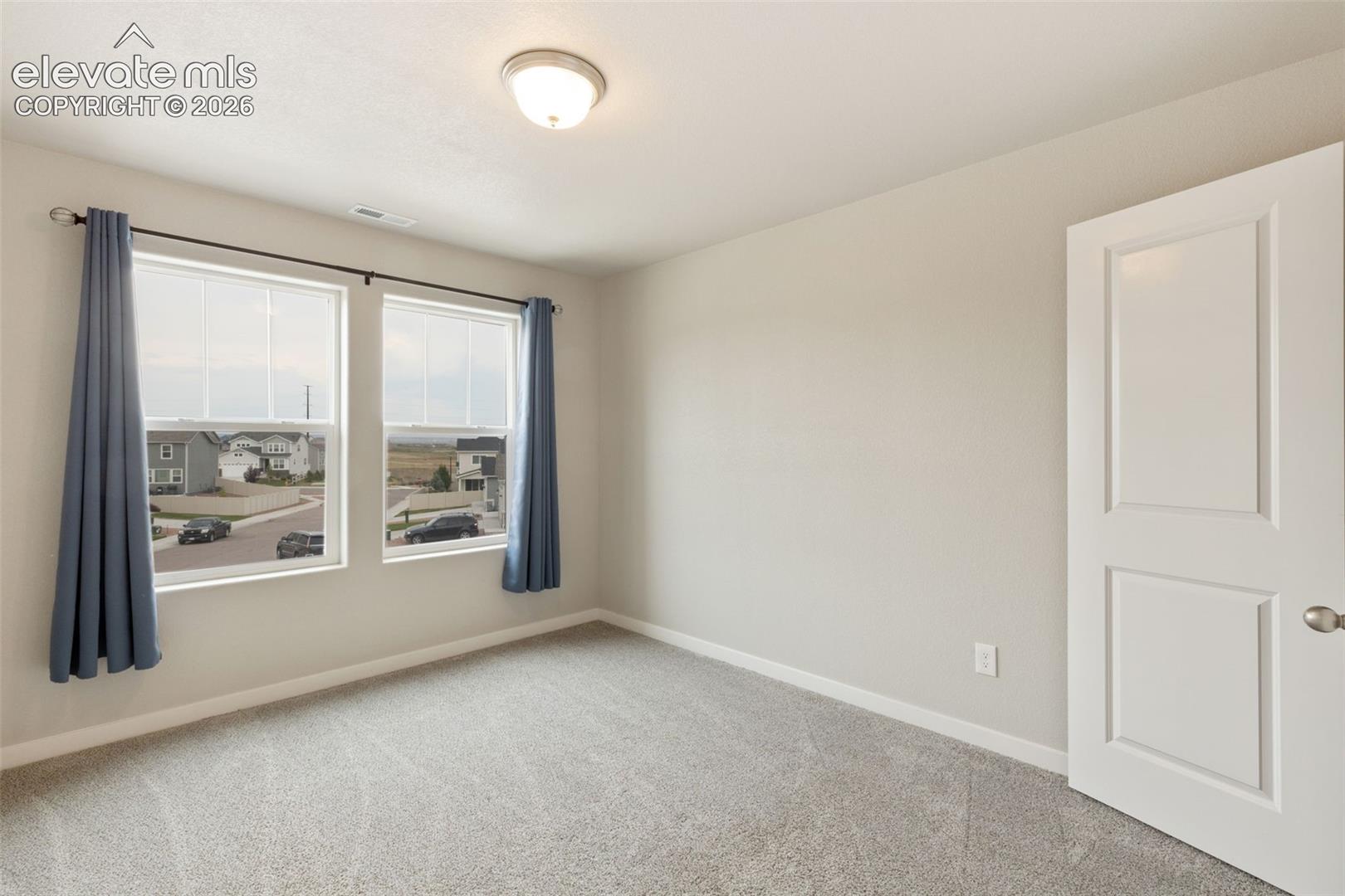 Bright room featuring two large windows with white blinds, grey carpet flooring, light neutral wall paint, and a two-panel interior door with brushed nickel hardware