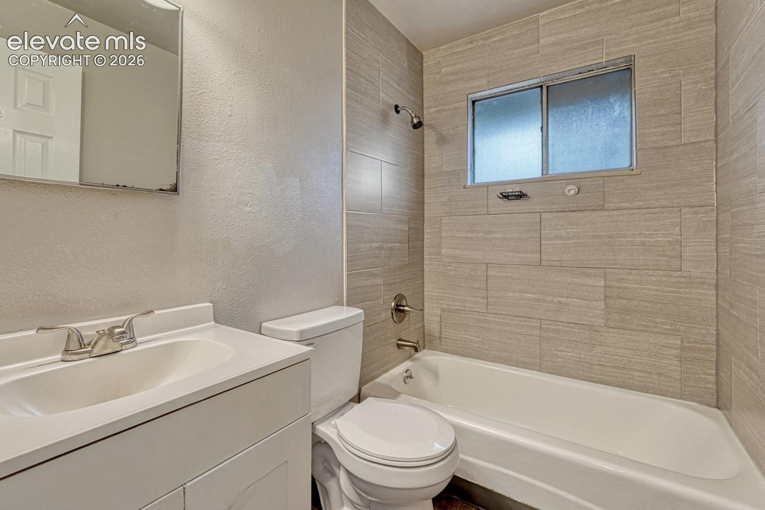 White vanity with integrated sink, chrome faucet, white toilet, tiled tub surround with recessed window, and a white bathtub