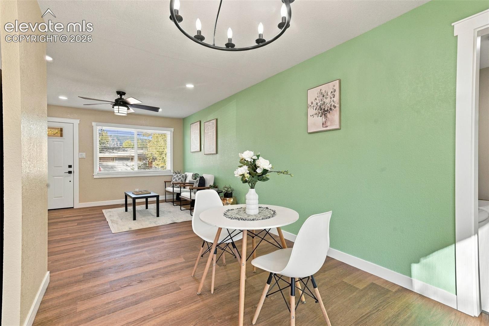 Open-concept living space featuring wood-finish flooring, a multi-arm chandelier, a ceiling fan, and a single-hung window with white casing