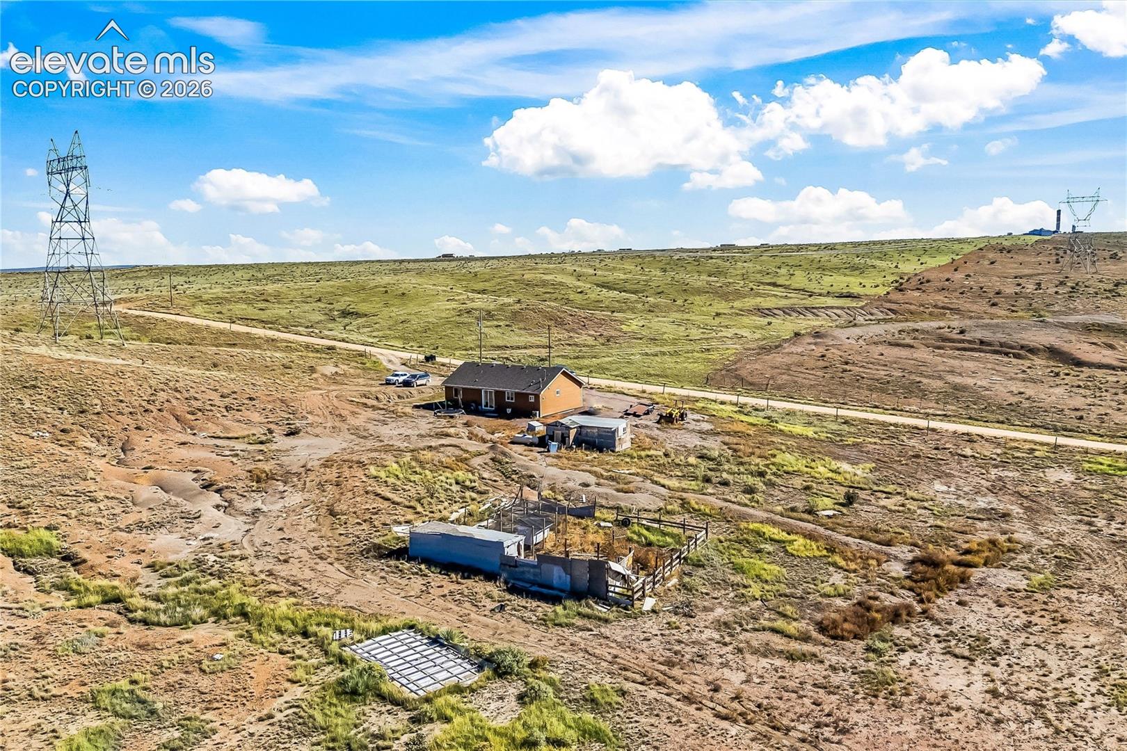 Aerial perspective of a detached residence situated on an expansive lot featuring natural desert terrain, a designated fenced enclosure, and a gravel road surface
