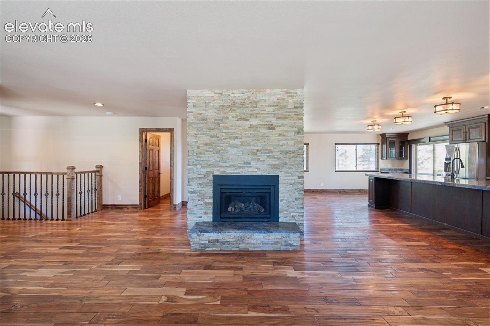 Floor-to-ceiling stone fireplace, wood-finish flooring, dark wood cabinetry with quartz-style countertops, and wrought-iron stair railing