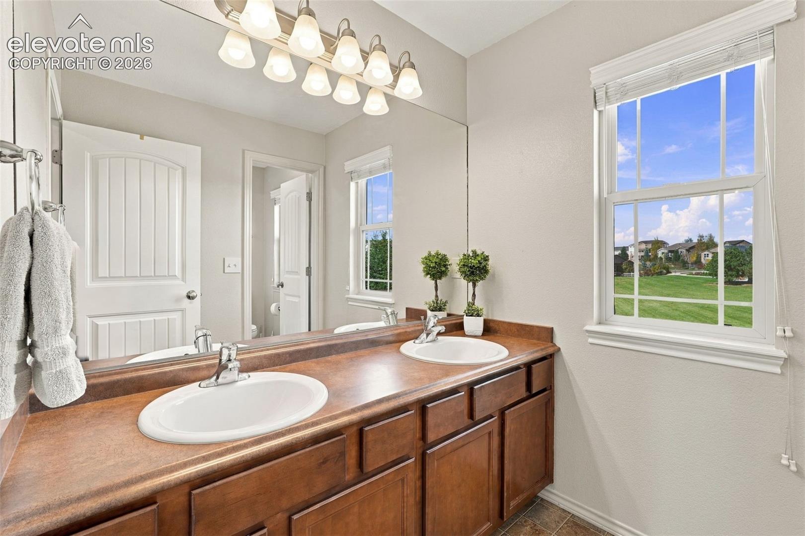 Double vanity featuring wood cabinetry, integrated dual sinks, a large wall-mounted mirror, and multi-bulb overhead lighting, complemented by a single-hung window
