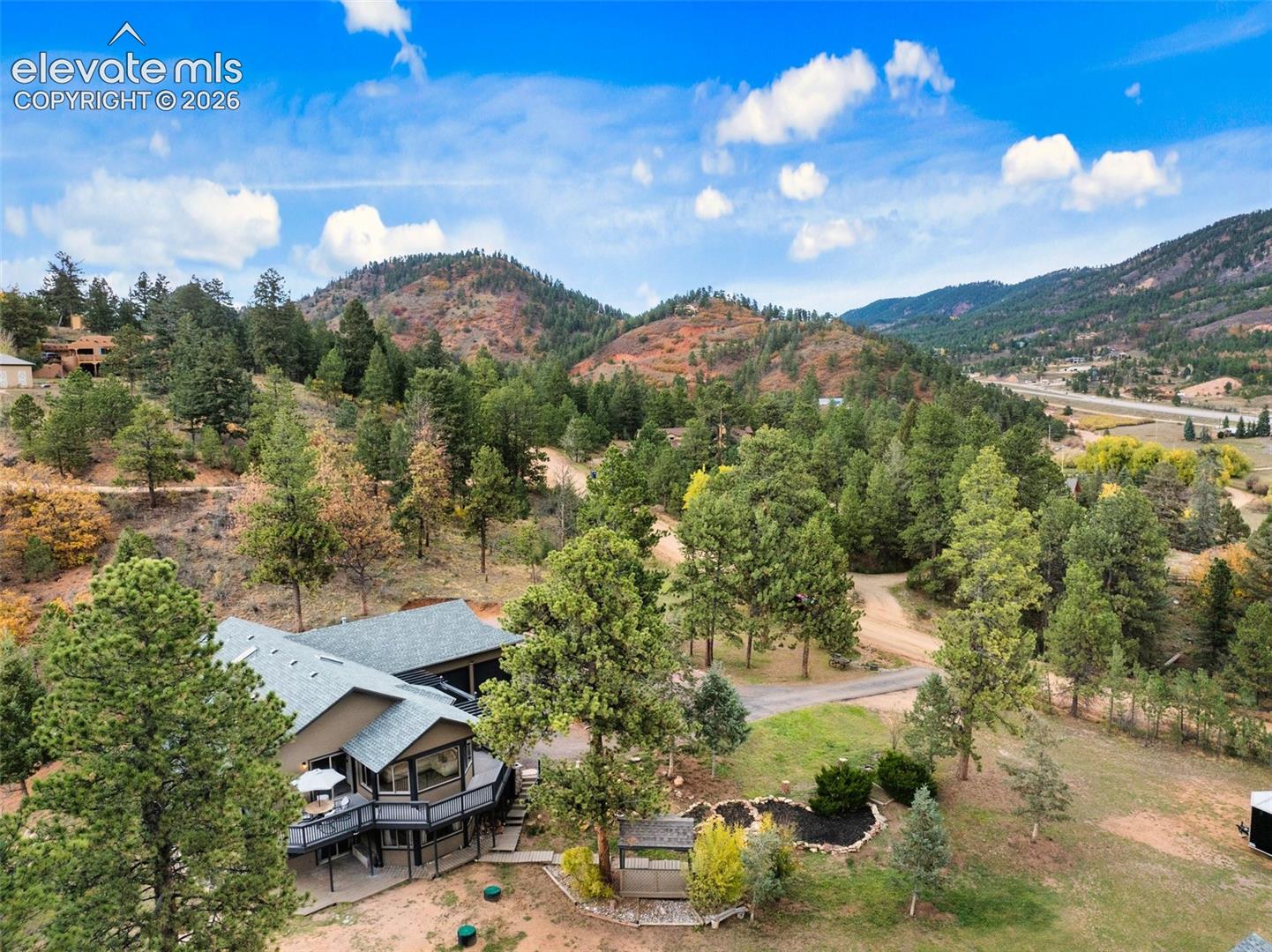 Aerial view of a multi-level residence featuring a wrap-around deck, shingle roofing, and extensive forested acreage set against a mountain backdrop