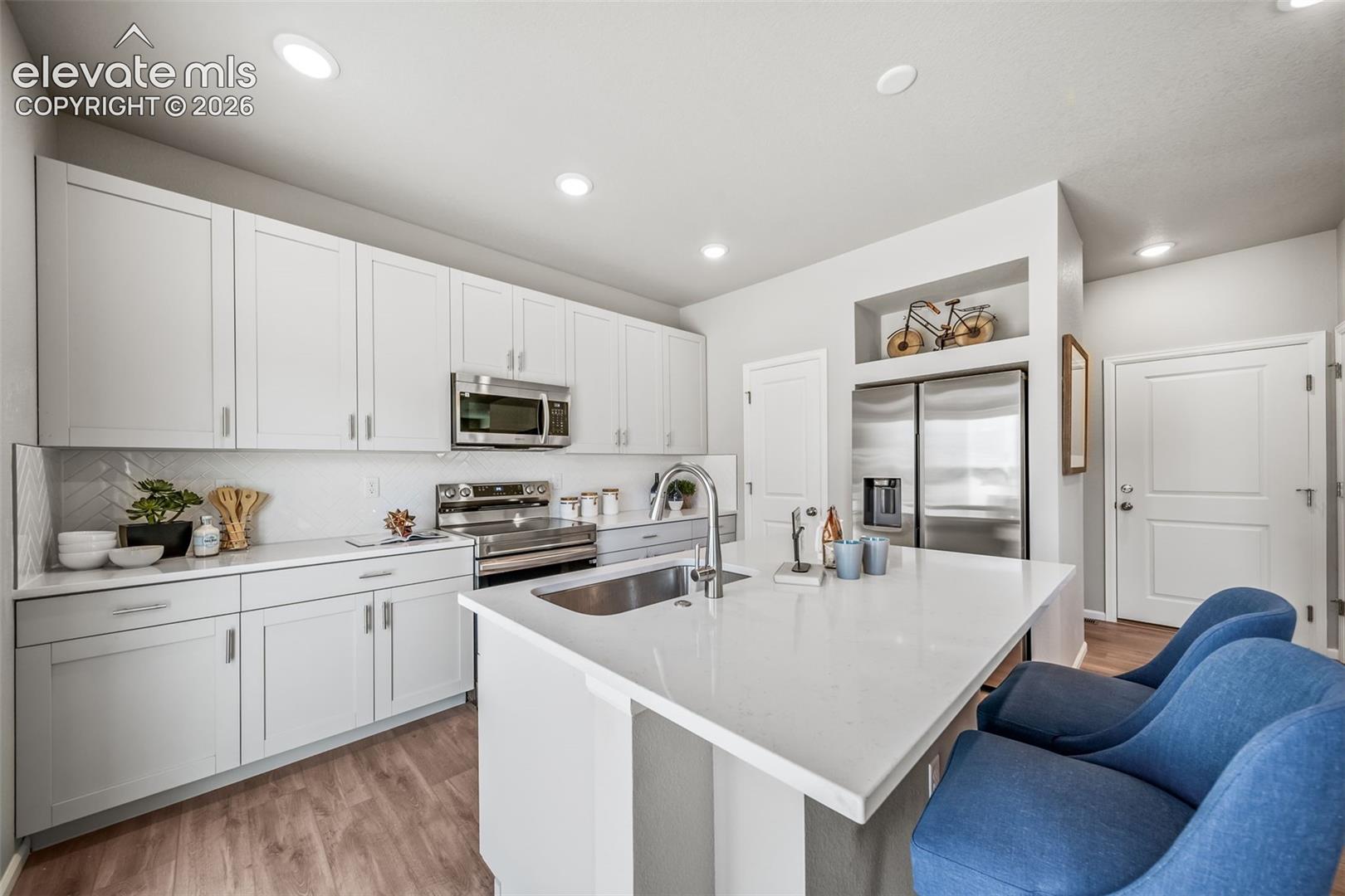 Kitchen featuring white shaker cabinetry, stainless steel appliances, a white island with an undermount sink, and wood-finish flooring
*photos of a model home*