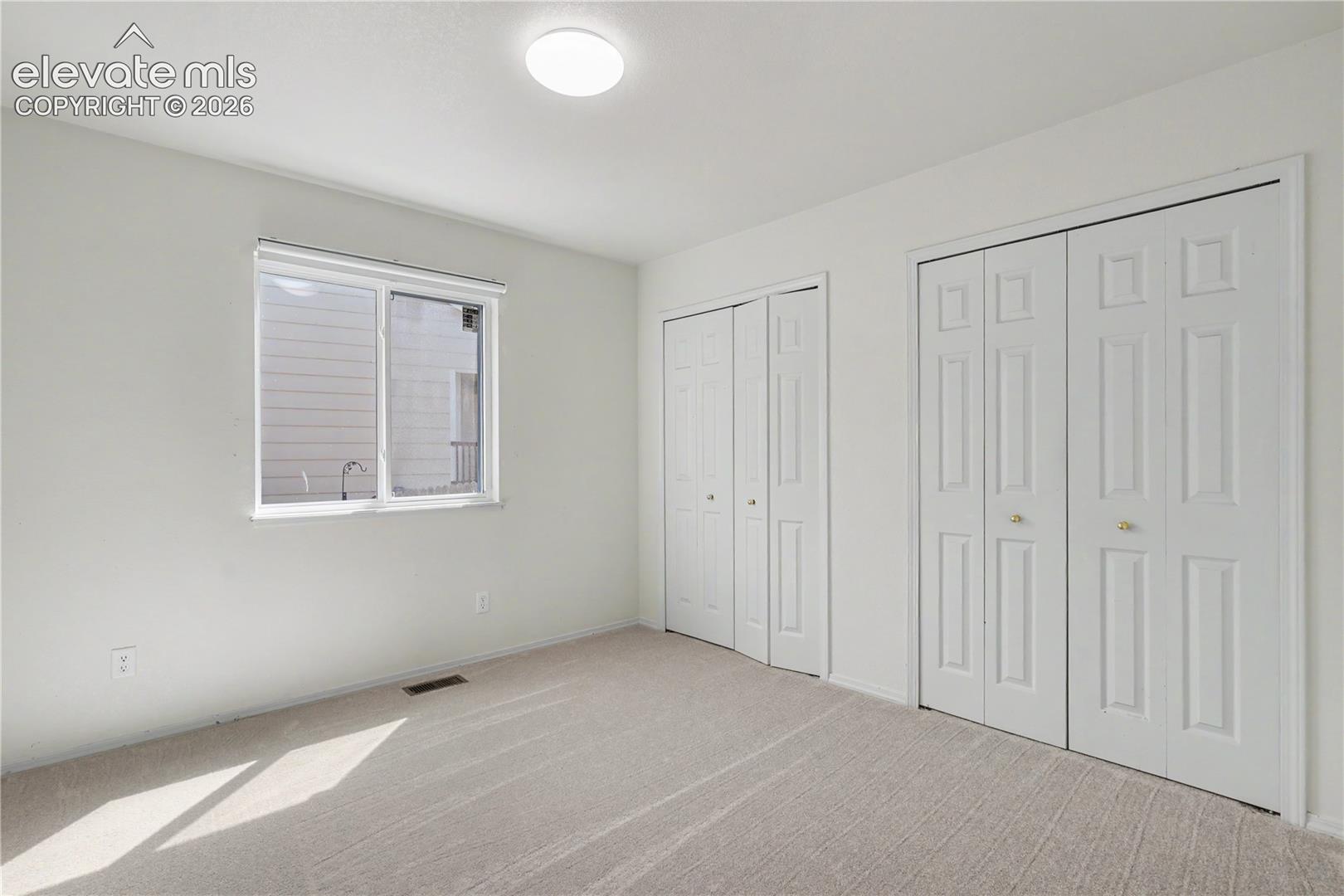 Neutral-toned carpeting extends throughout this interior space, featuring a window with white trim, a flush-mount ceiling light fixture, and two sets of bi-fold closet doors with brass-toned hardware