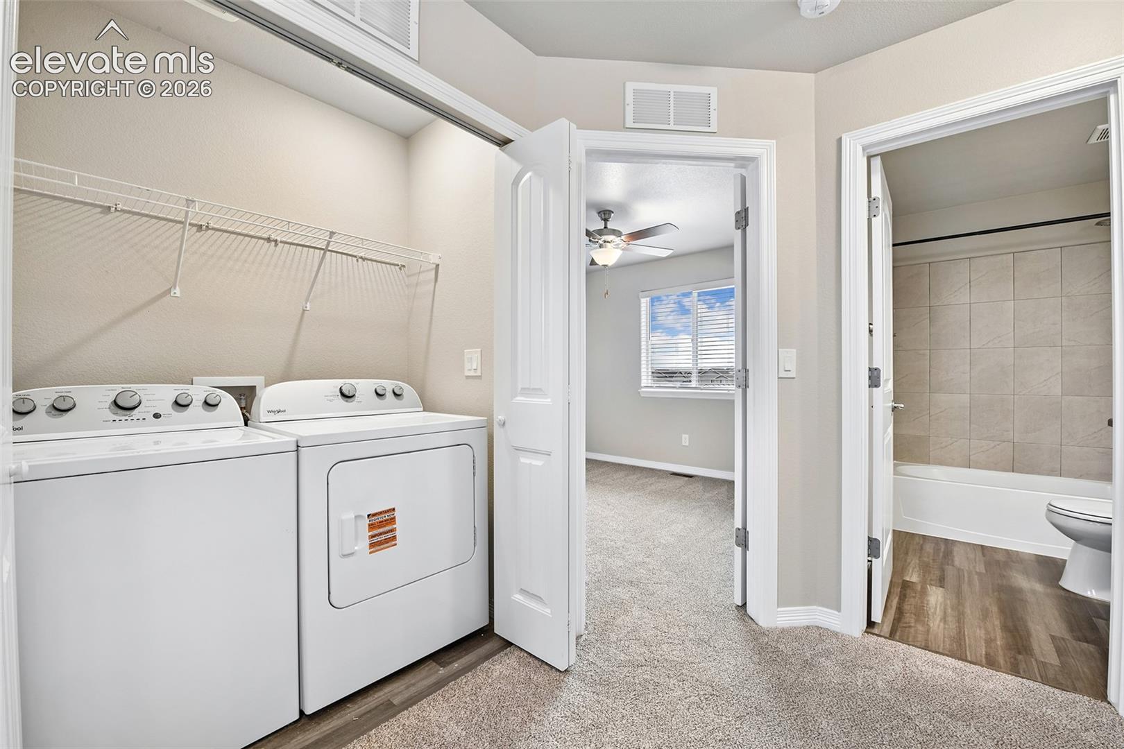 Laundry area featuring wire shelving, wall-to-wall carpeting, and neutral paint, with access to a bathroom finished with wood-finish flooring and tiled tub surround