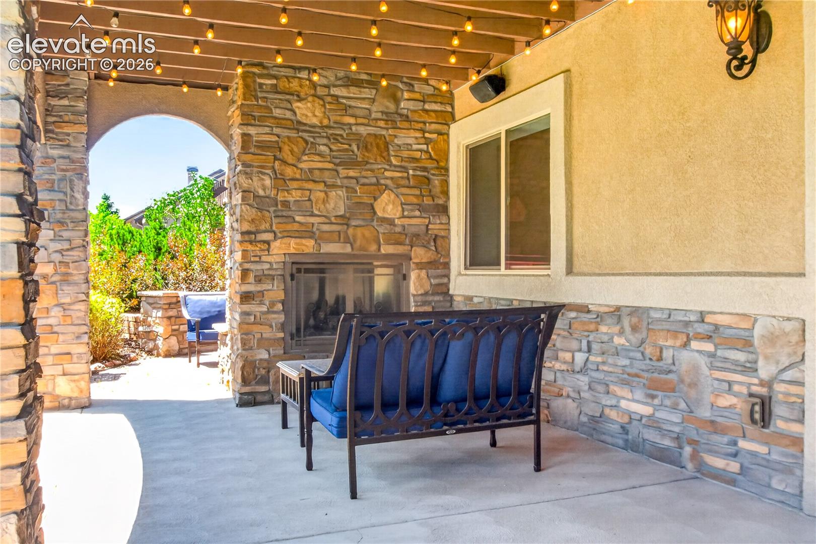 Covered patio featuring a stone-clad exterior wall, built-in outdoor fireplace, concrete flooring, arched passageway, and overhead wood beam ceiling structure