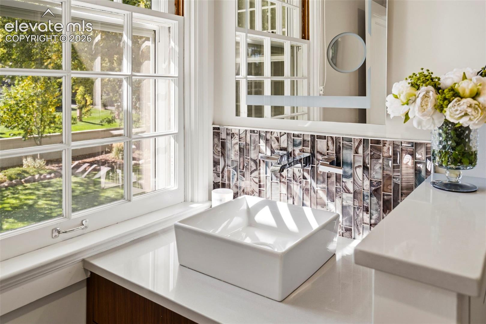 White square vessel sink with wall-mounted faucet, stone surface countertop, decorative glass mosaic backsplash, and muntin-style window