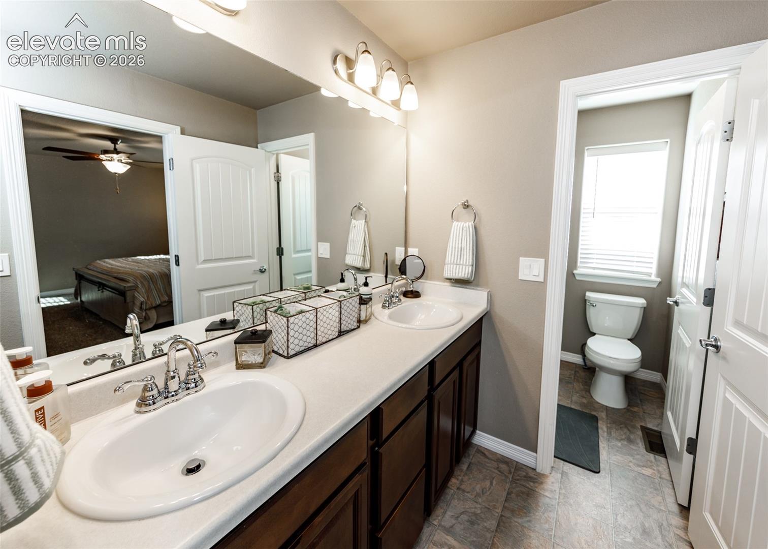 Dual-sink vanity with cabinetry, solid-surface countertops, chrome finish hardware, wall-mounted lighting, and a private water closet