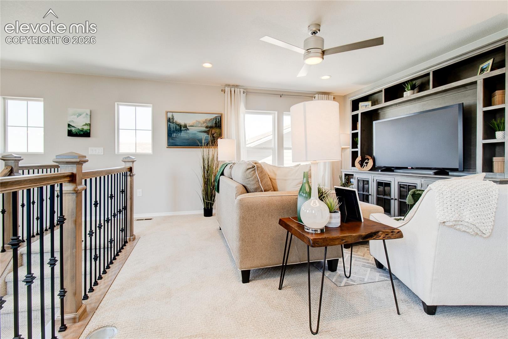 Custom built-in media cabinetry with display shelving, metal spindle staircase railing, ceiling fan with integrated lighting, and neutral carpeting