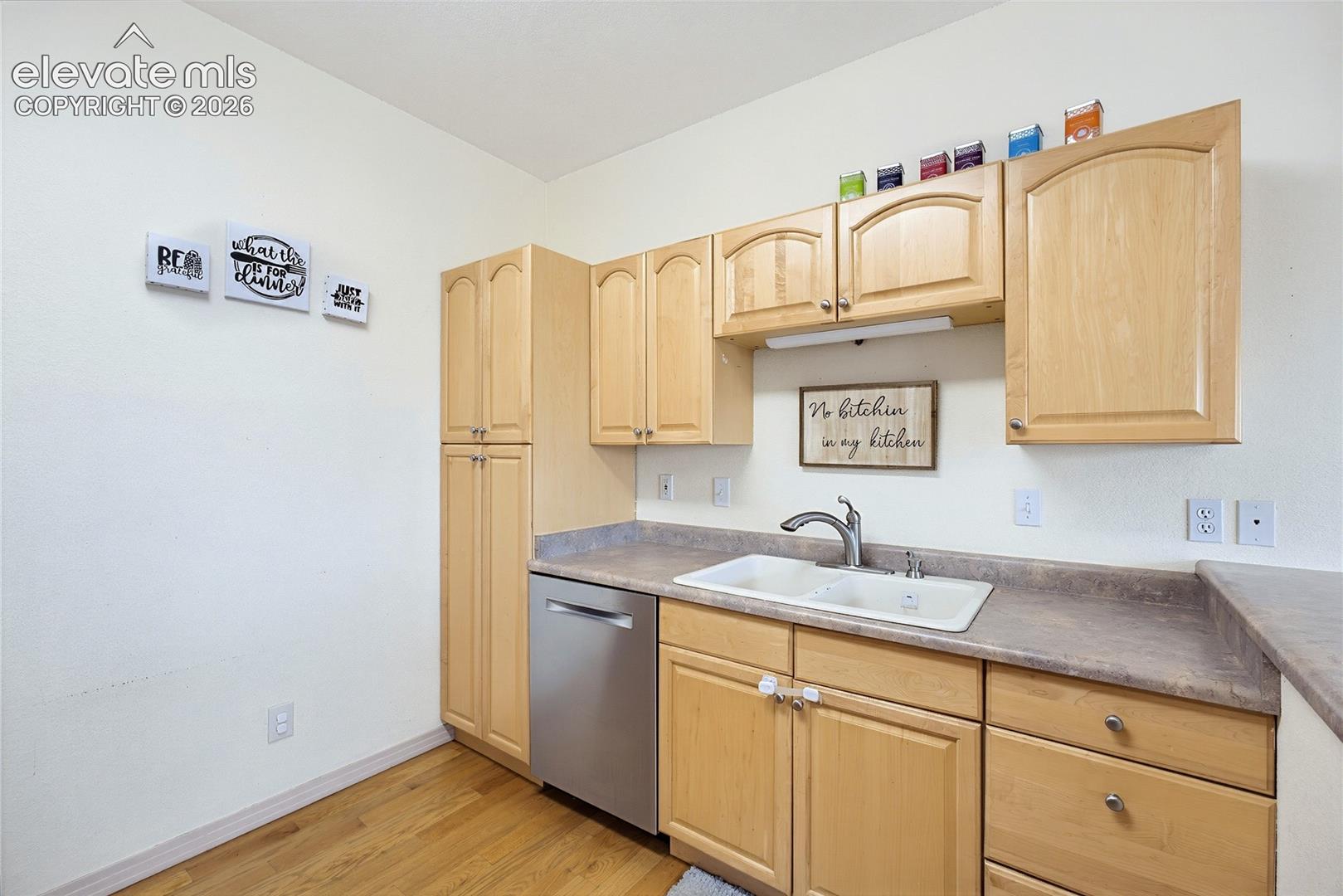 Light wood-finish cabinetry with silver hardware, dual-basin sink with a chrome faucet, stainless steel dishwasher, and wood-finish flooring