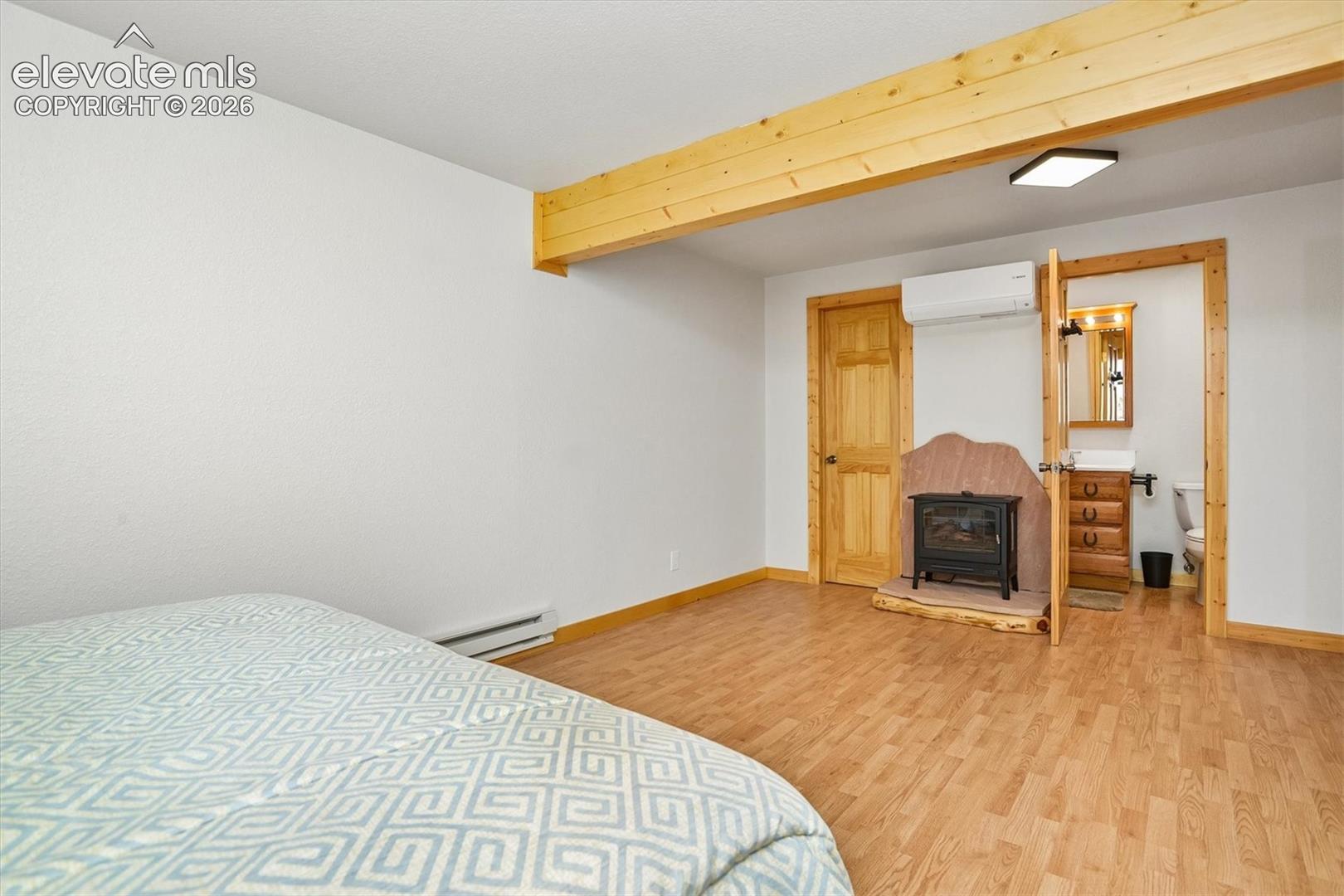 Spacious room featuring wood-finish flooring, exposed wood beam, a freestanding electric fireplace with a stone surround, and an en-suite half bathroom with a wood vanity and mirror
