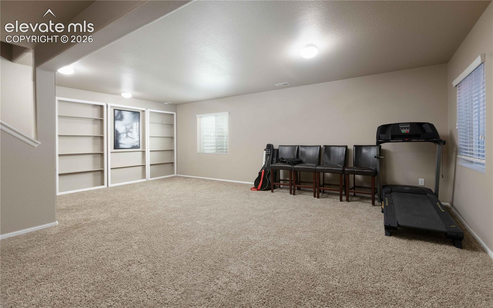 Neutral-toned interior space featuring wall-to-wall carpeting, recessed lighting, white built-in shelving, and two windows with horizontal blinds