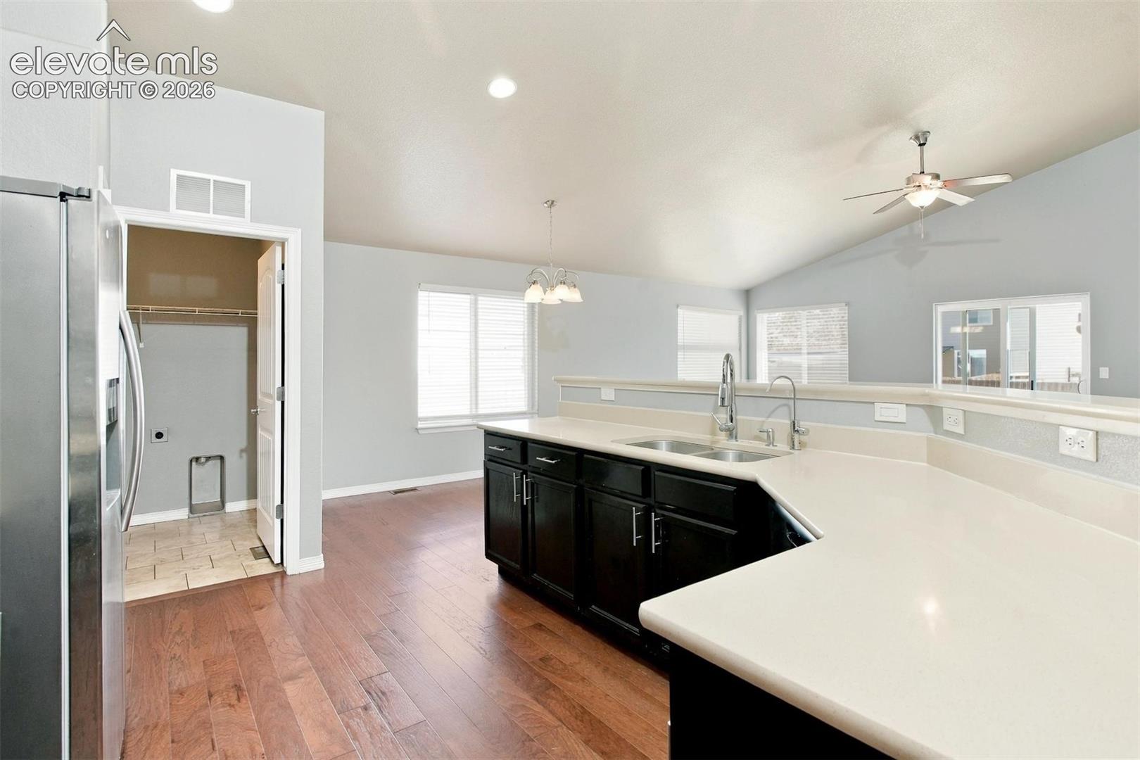 Kitchen featuring dark cabinetry, expansive solid-surface countertops, dual-basin stainless steel sink with a high-arch pull-down faucet, wood-finish flooring, and a vaulted ceiling