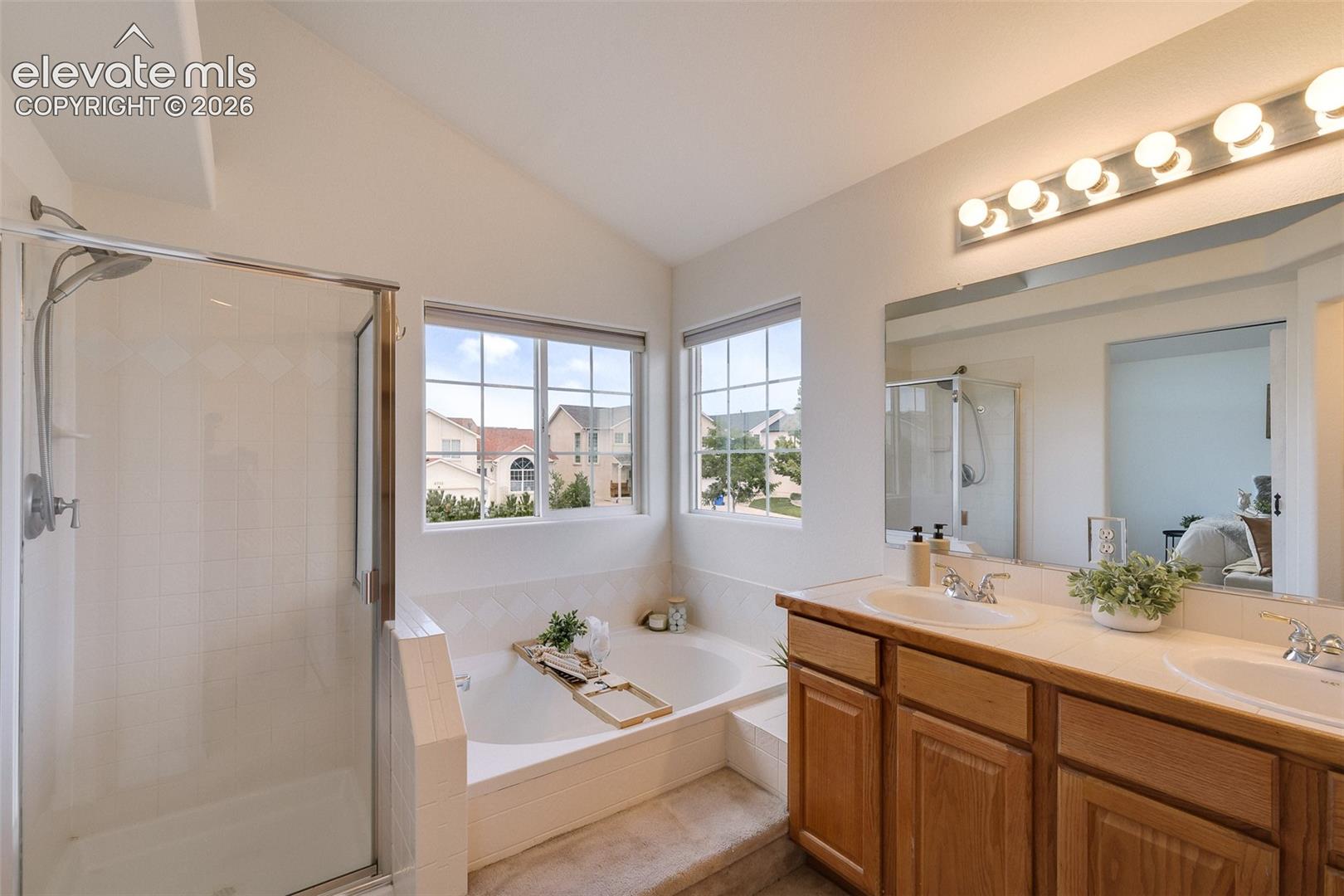 Dual windows, vaulted ceiling, glass-enclosed shower, built-in bathtub, and a dual vanity with wood-finish cabinetry