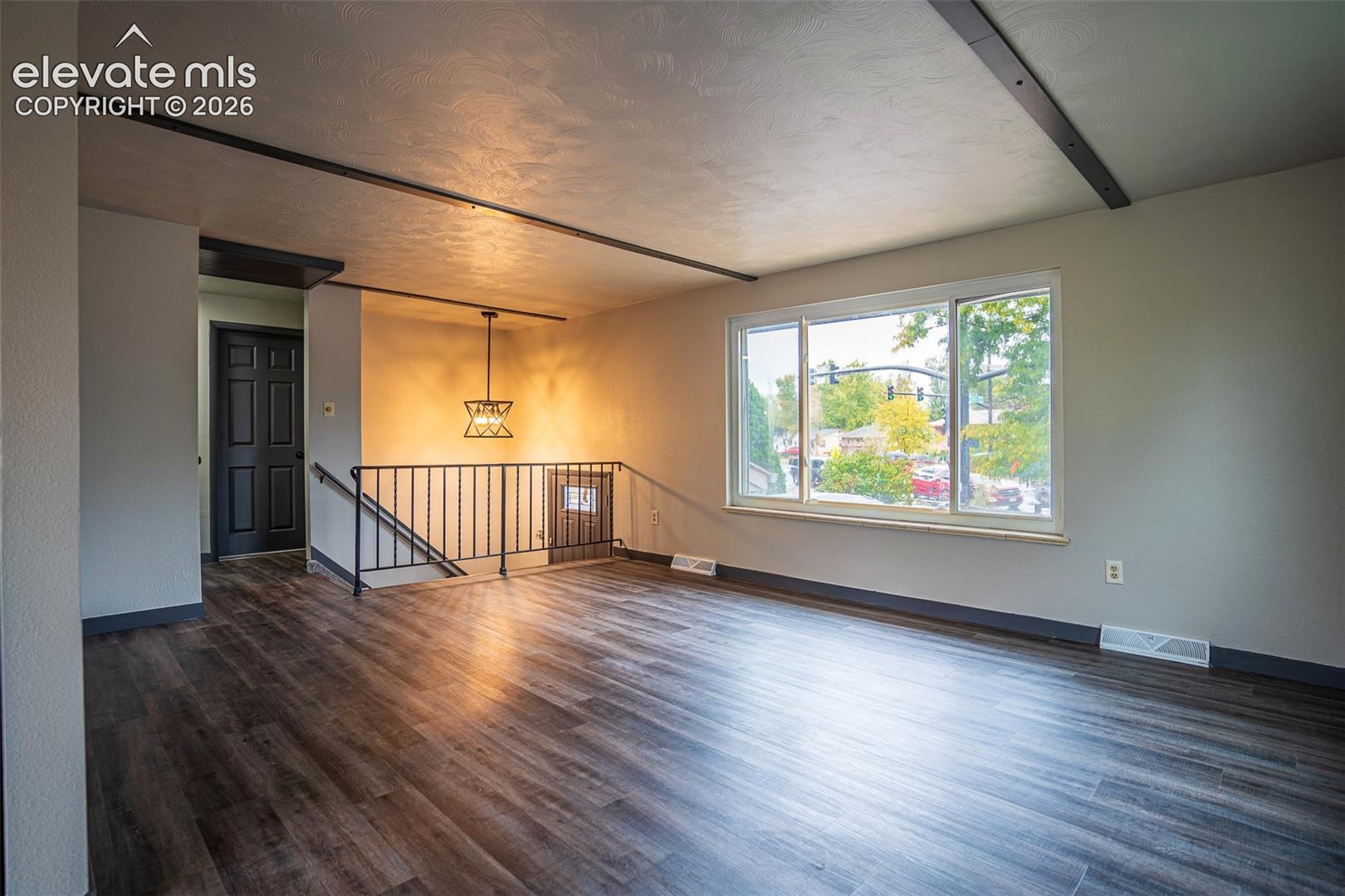 Open living space featuring wood-finish flooring, exposed structural ceiling beams, a large picture window, a geometric pendant light fixture, and wrought iron staircase railing