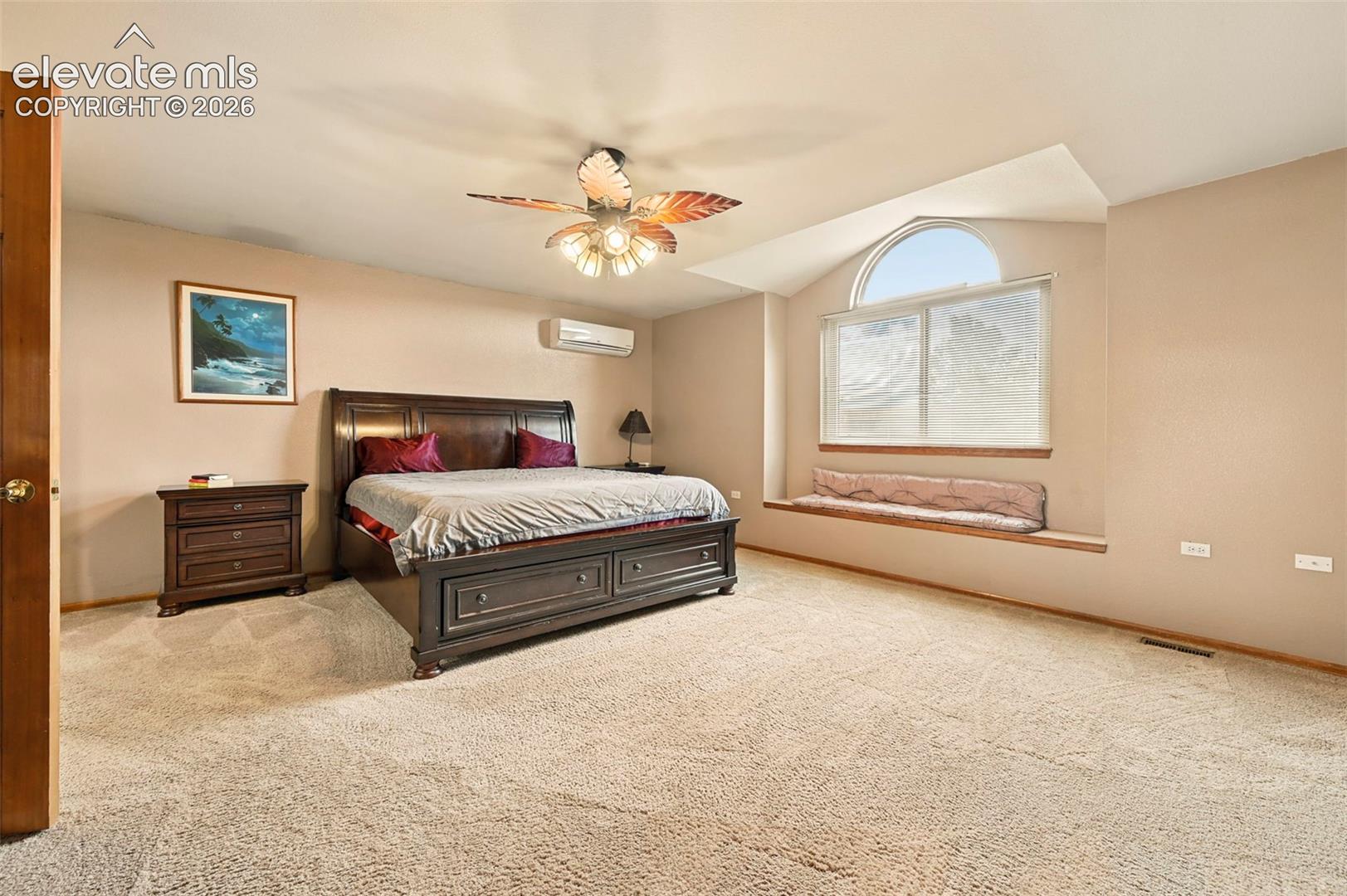 Primary bedroom with ceiling fan, arched window, window bench, wall-mounted climate control unit, and carpeted flooring
