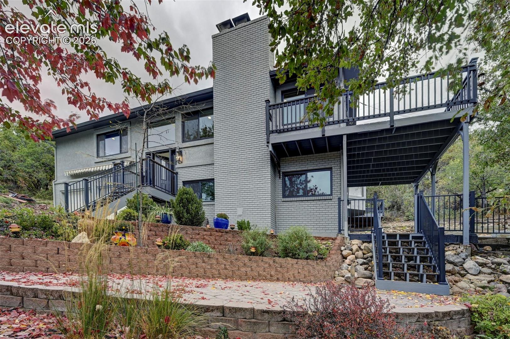 Multi-level home exterior featuring a painted brick facade, a central masonry chimney, an elevated wooden deck with metal railing, and terraced stone retaining walls