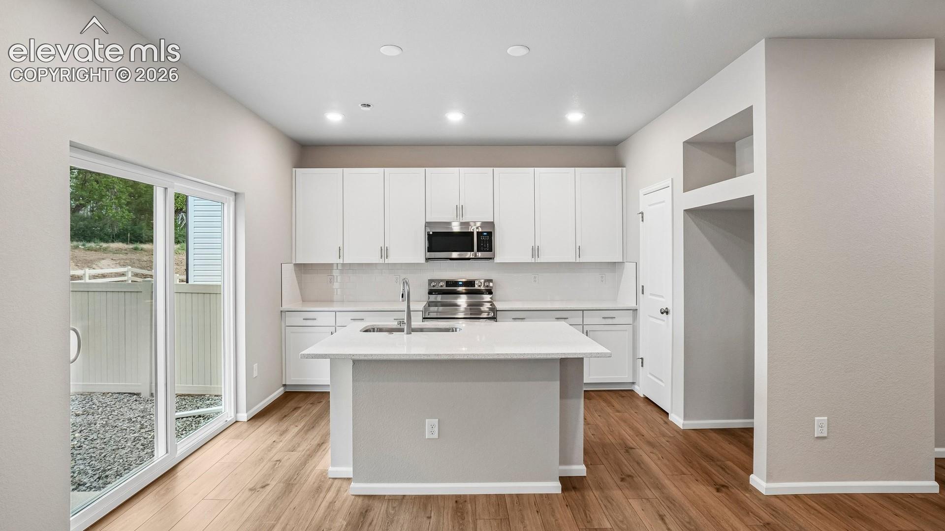 Kitchen featuring modern white cabinetry, stainless steel appliances, a center island with sink, and wood-finish flooring
*photos of a model home*